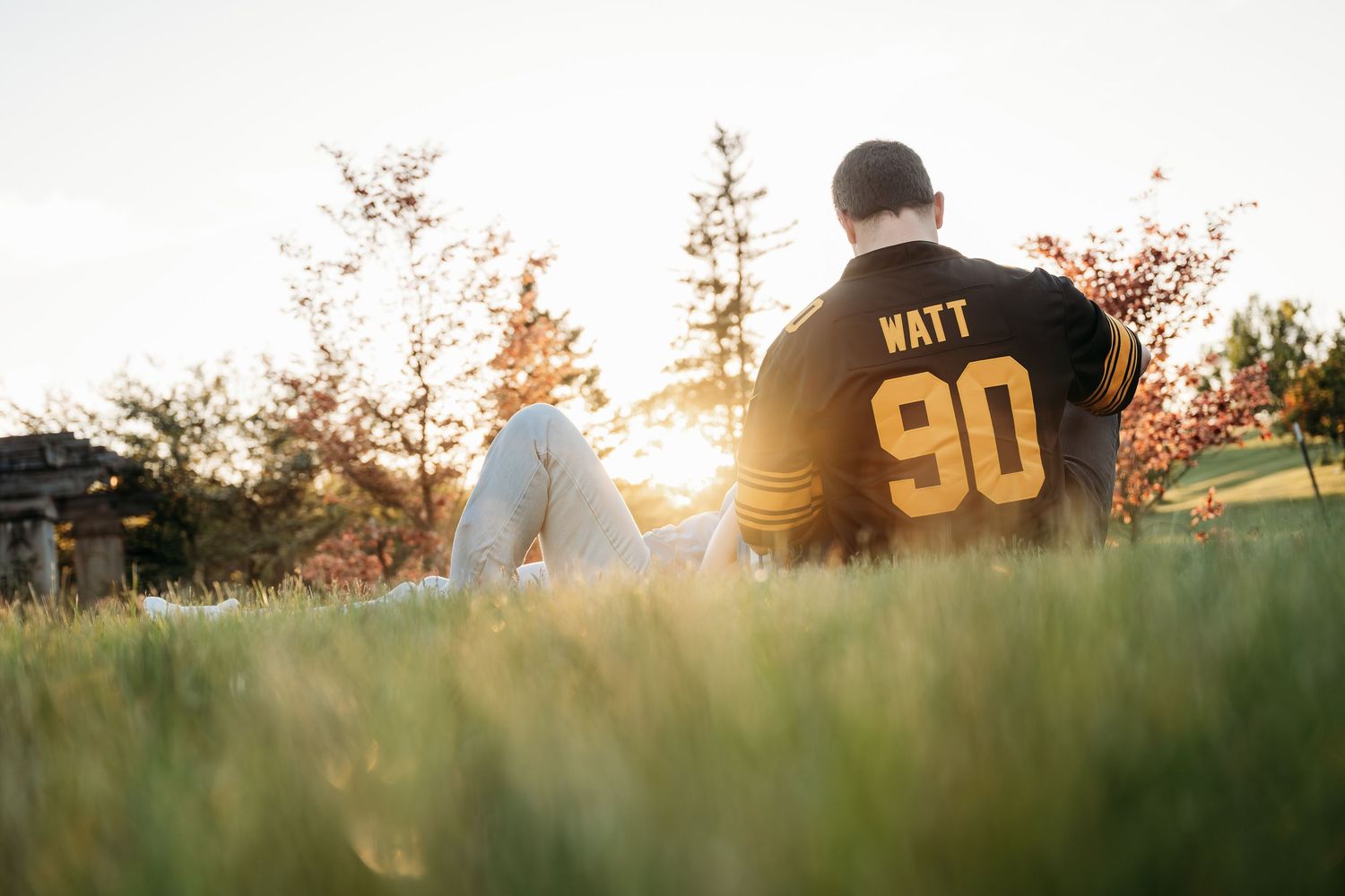 Person sitting on grass at sunset wearing a Watt #90 football jersey, viewed from behind.