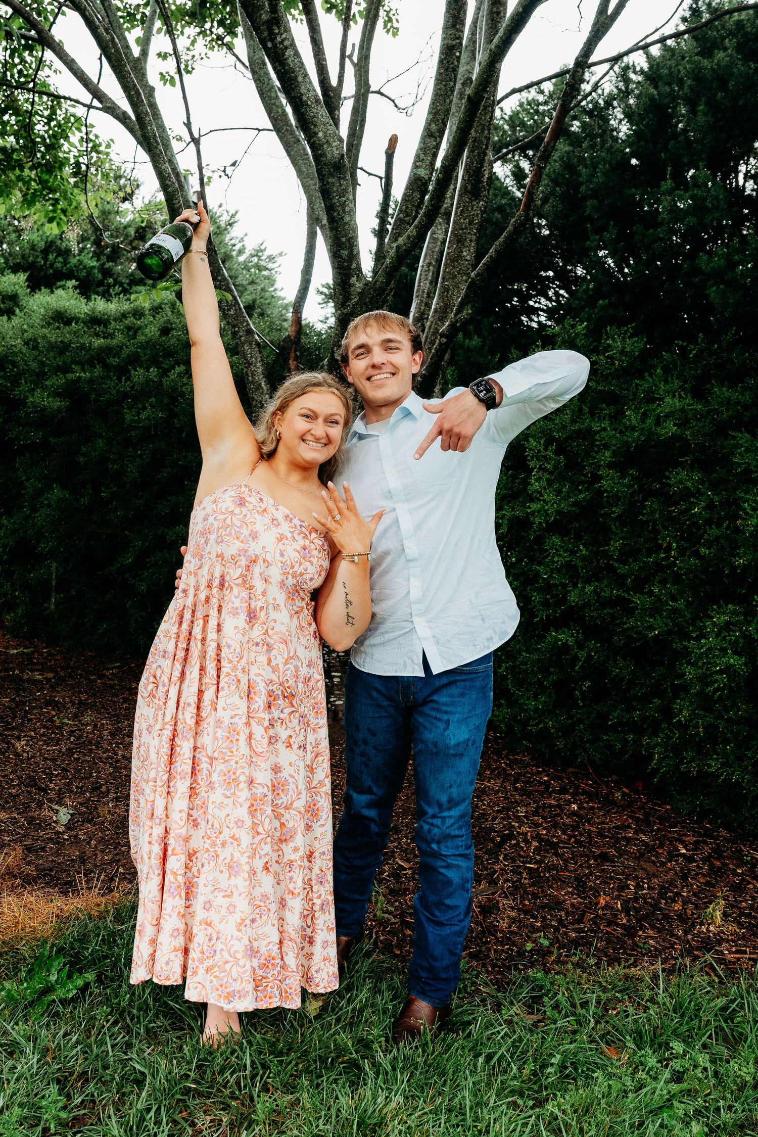 Happy couple celebrates engagement outdoors, woman showing ring while both pose joyfully among lush green trees.