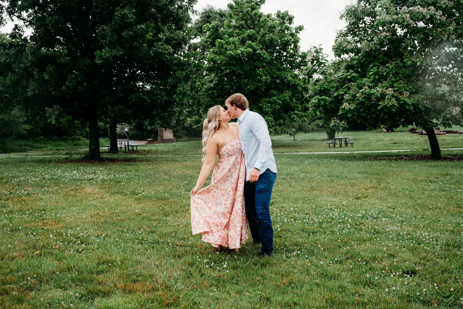 Couple sharing a romantic kiss in a lush green park, woman wearing a floral pink dress, surrounded by trees.