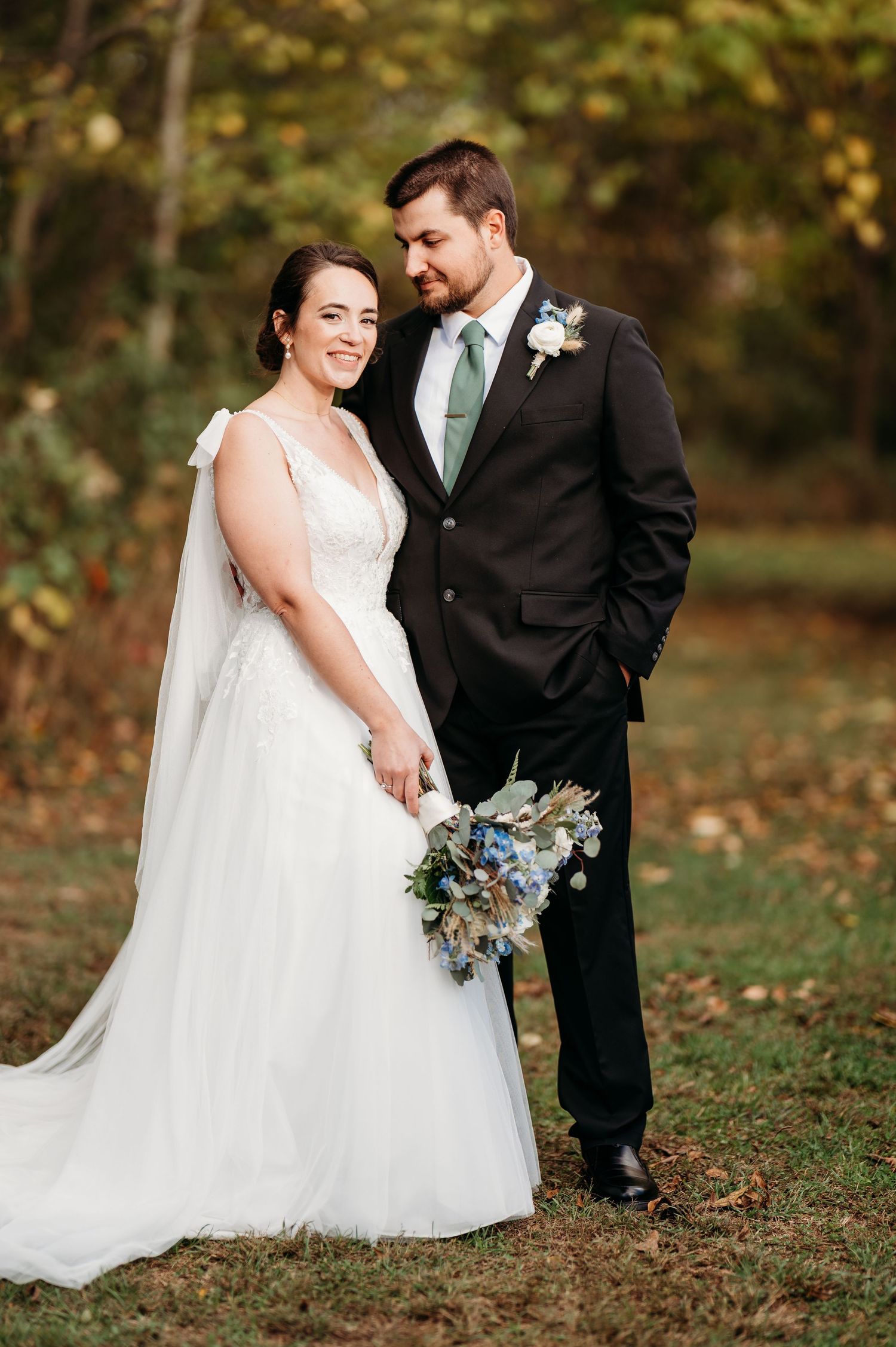 A couple in wedding attire poses outdoors in autumn, with the bride holding a blue and white floral bouquet.