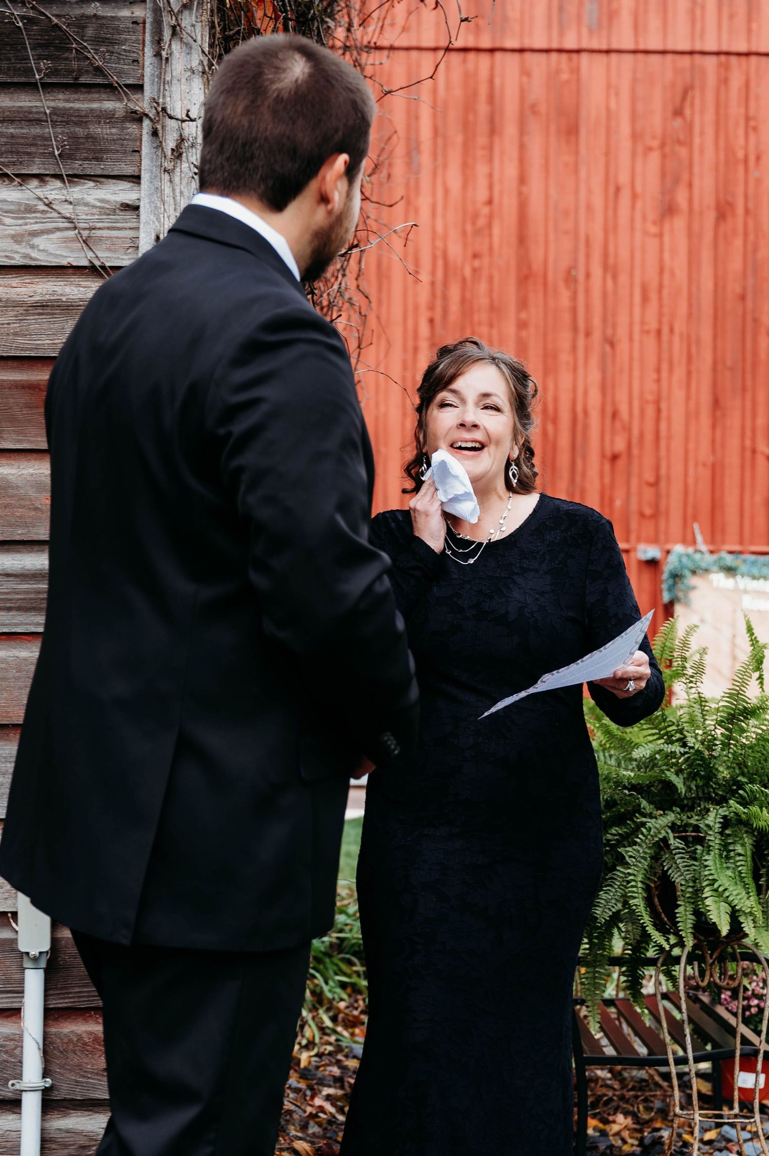 Two people in formal black attire share a joyful moment during a ceremony against a rustic red barn wall.