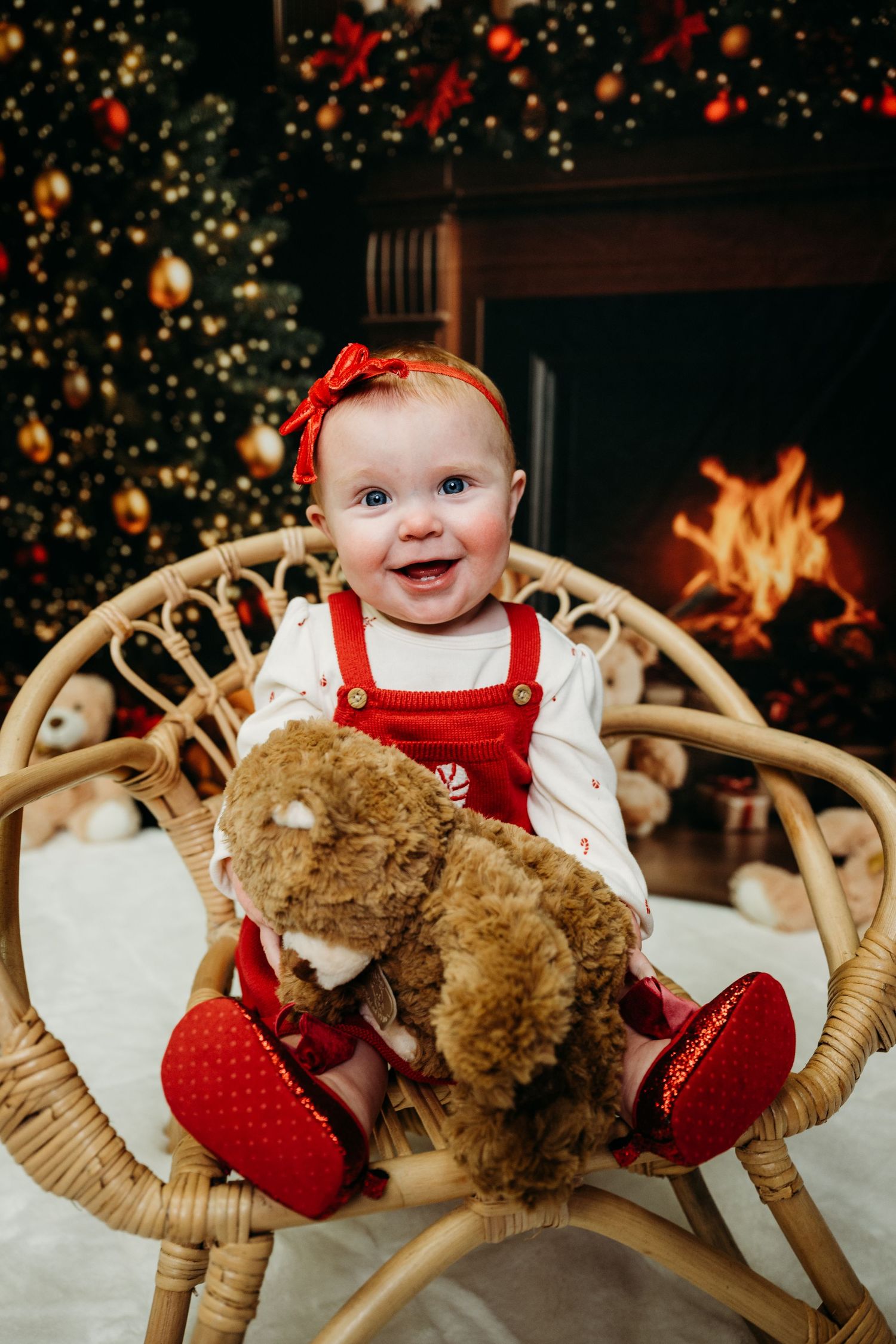 Baby in red outfit sitting in wicker chair with teddy bear and Christmas decorations.
