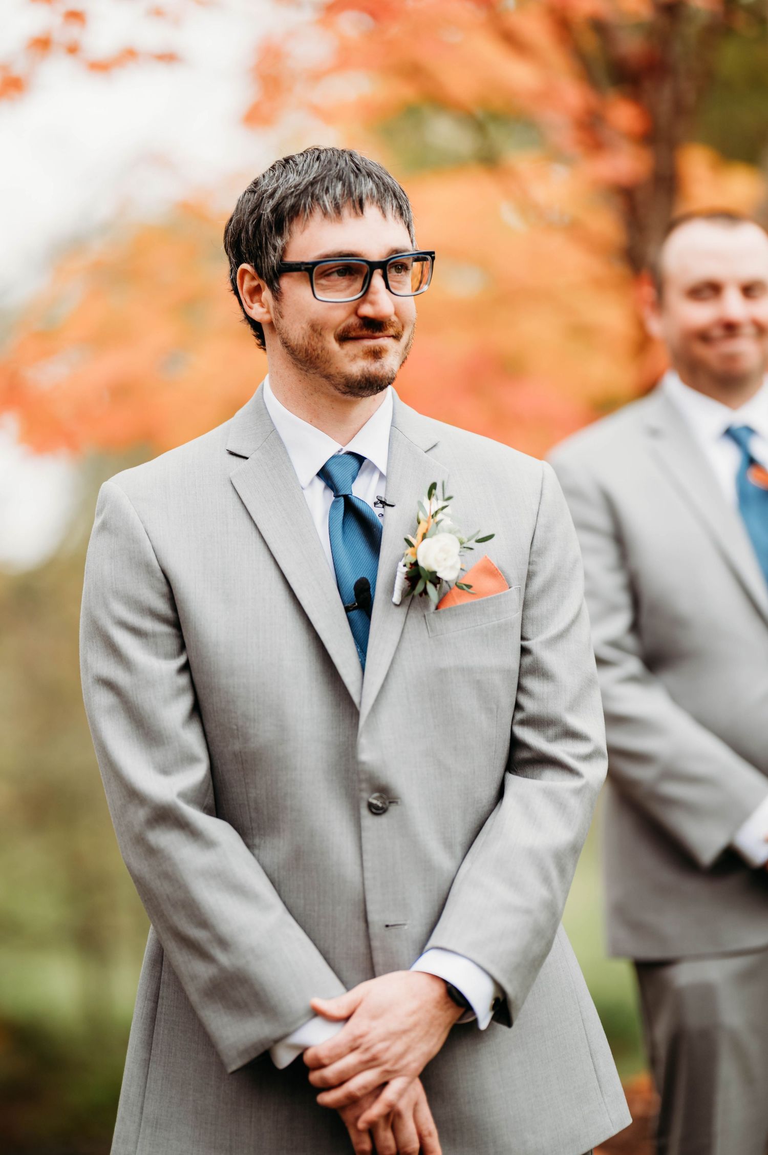 Wedding party members in gray suits and blue ties stand together during an autumn outdoor ceremony.