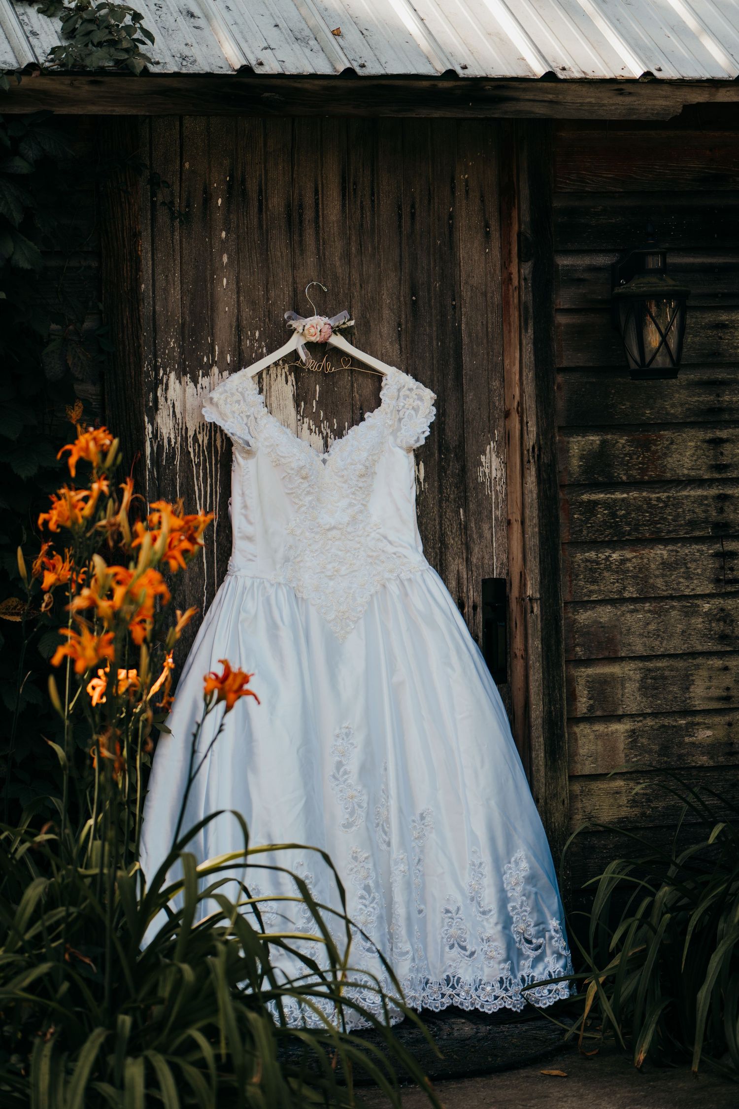 A white lace wedding dress hangs on a rustic wooden wall beside blooming orange day lilies.