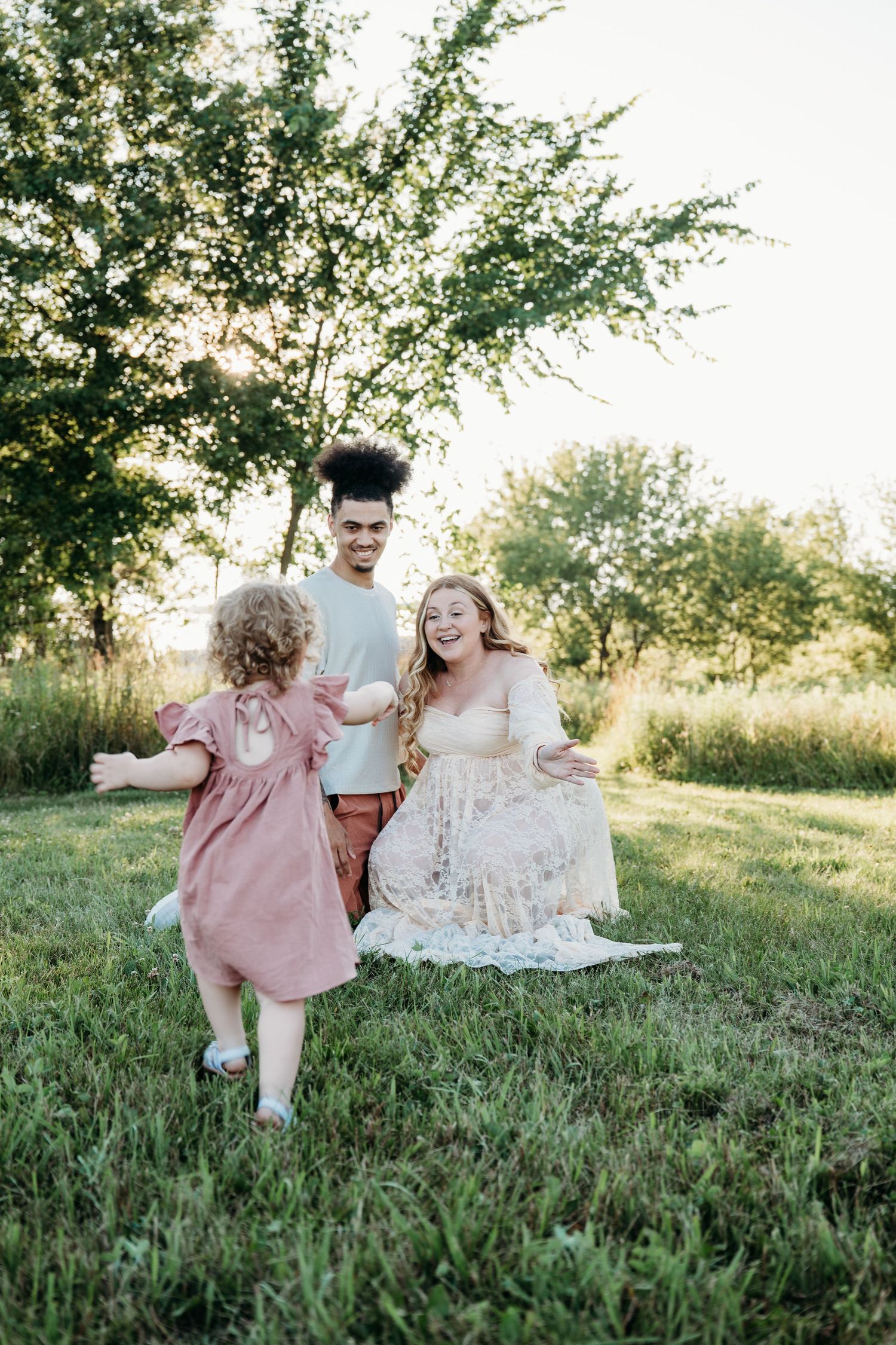 A candid outdoor family photo session featuring parents and a small child sitting together in a grass field at sunset.