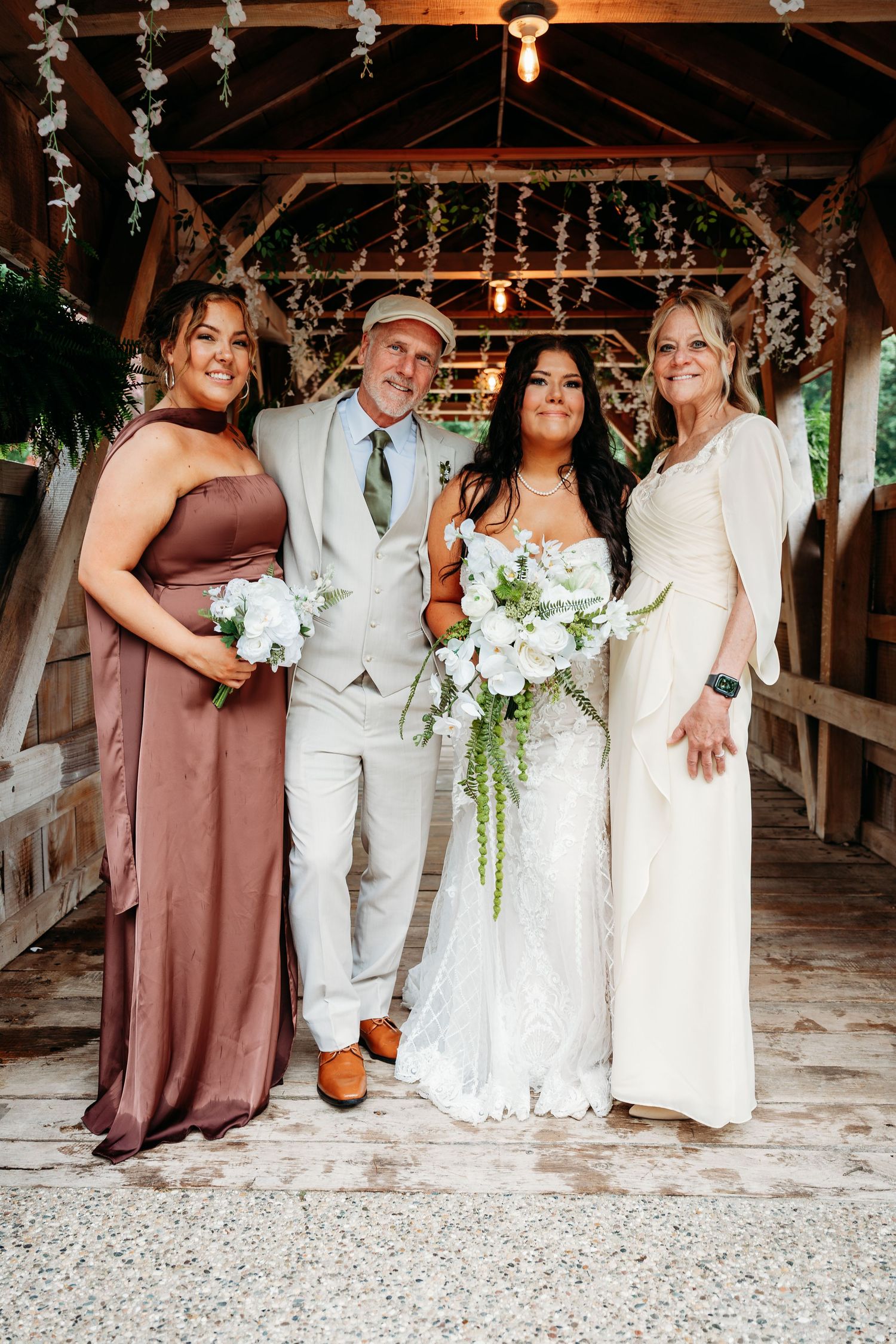 Bride in lace gown holds white bouquet posing with groom in gray suit, bridesmaid in mauve, and woman in ivory dress.