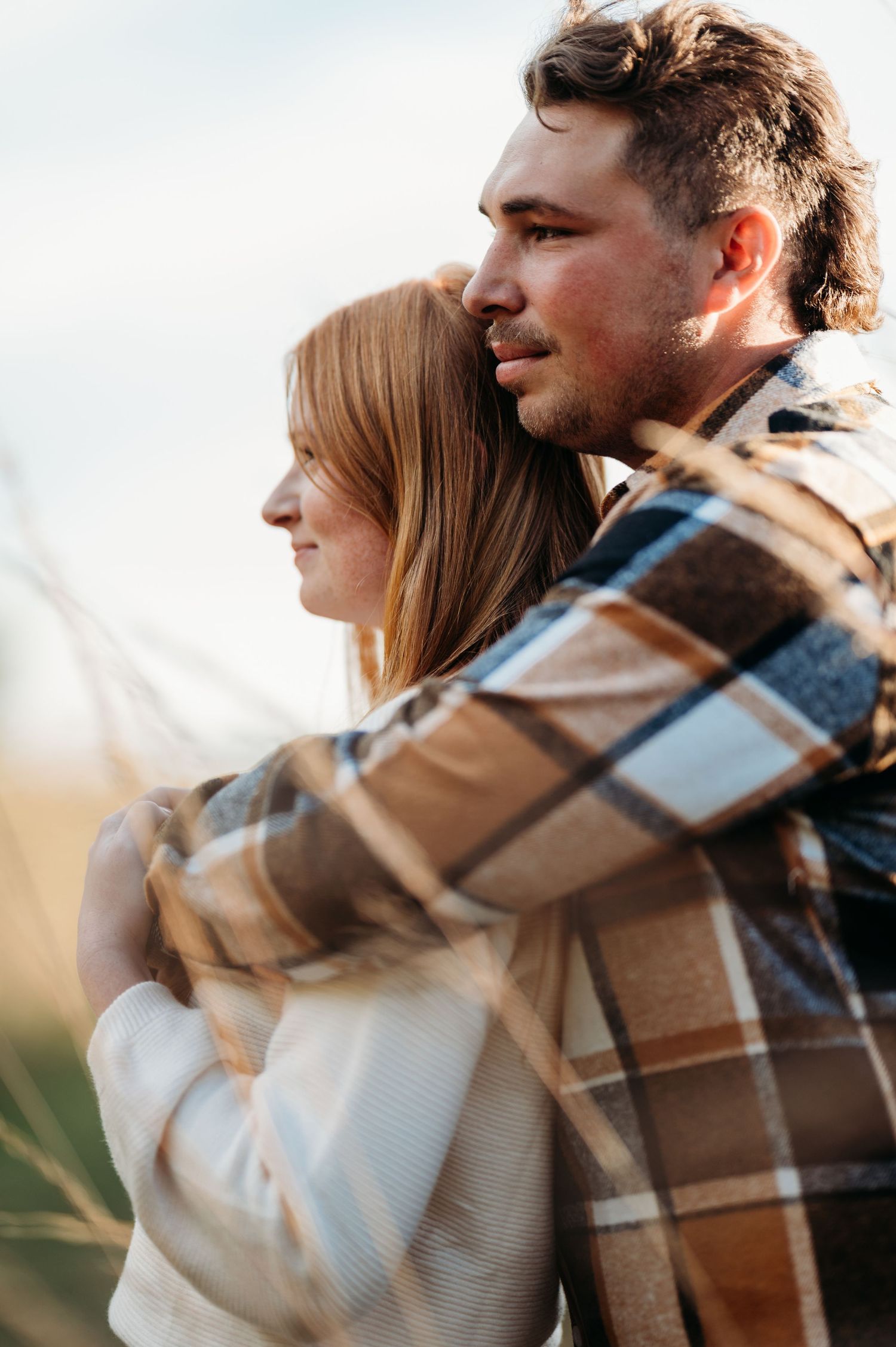A couple wrapped in a cozy plaid blanket share a tender embrace during a golden hour outdoor photoshoot.