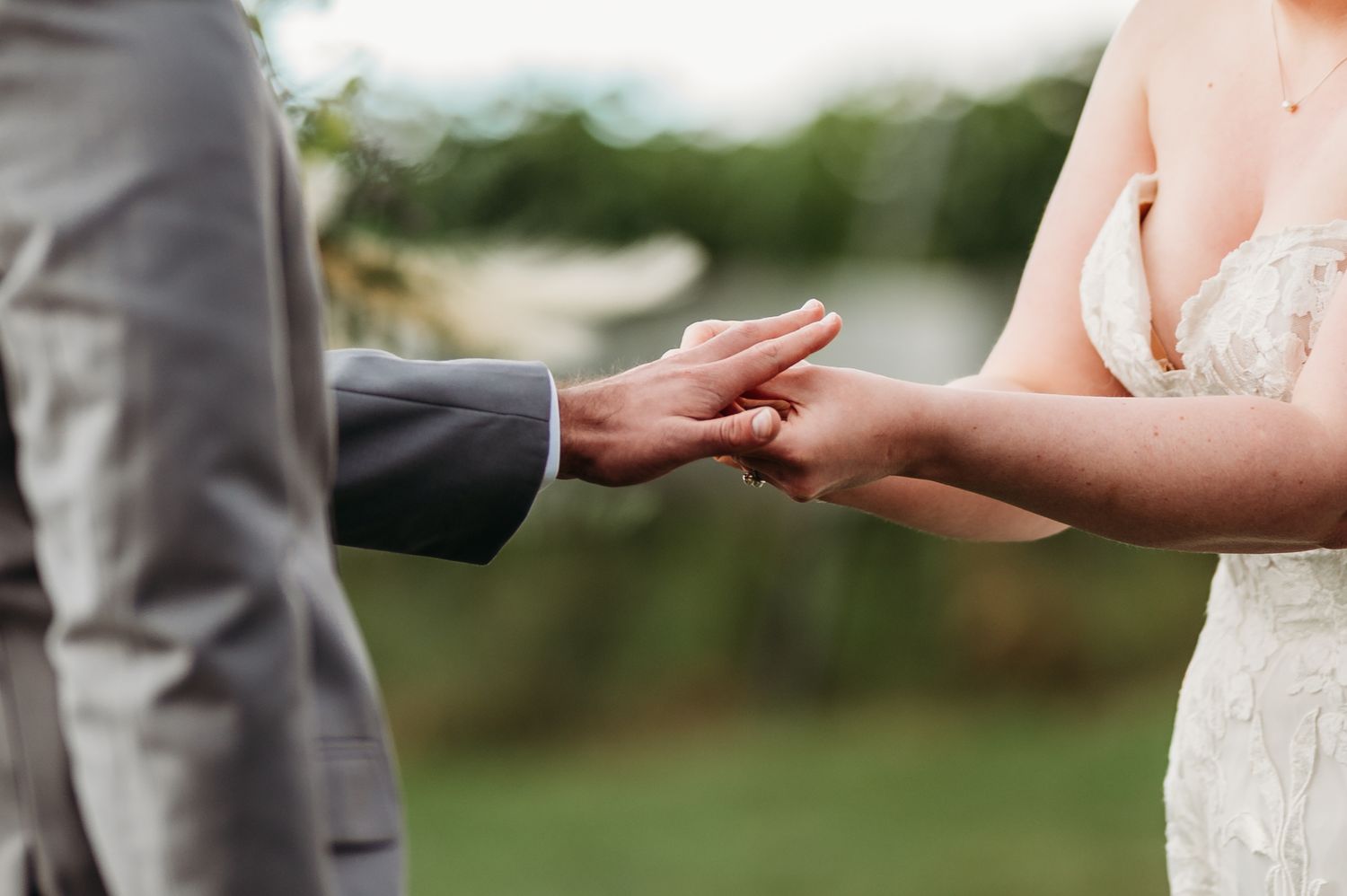 Close-up of hands exchanging wedding rings during an outdoor ceremony with lace dress and gray suit details.