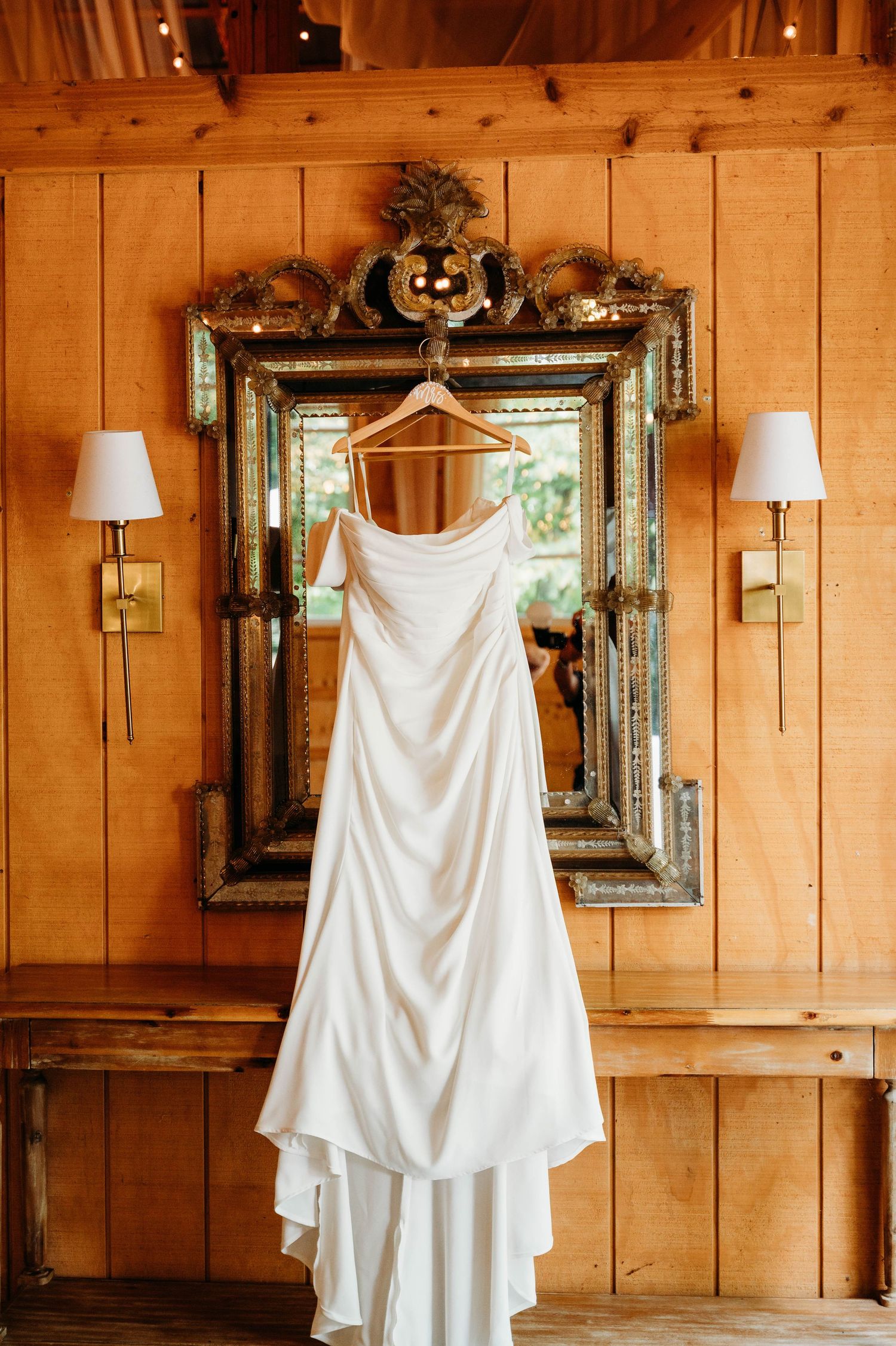 White wedding dress hangs on wooden hanger against rustic wooden wall with decorative mirrors and decor.