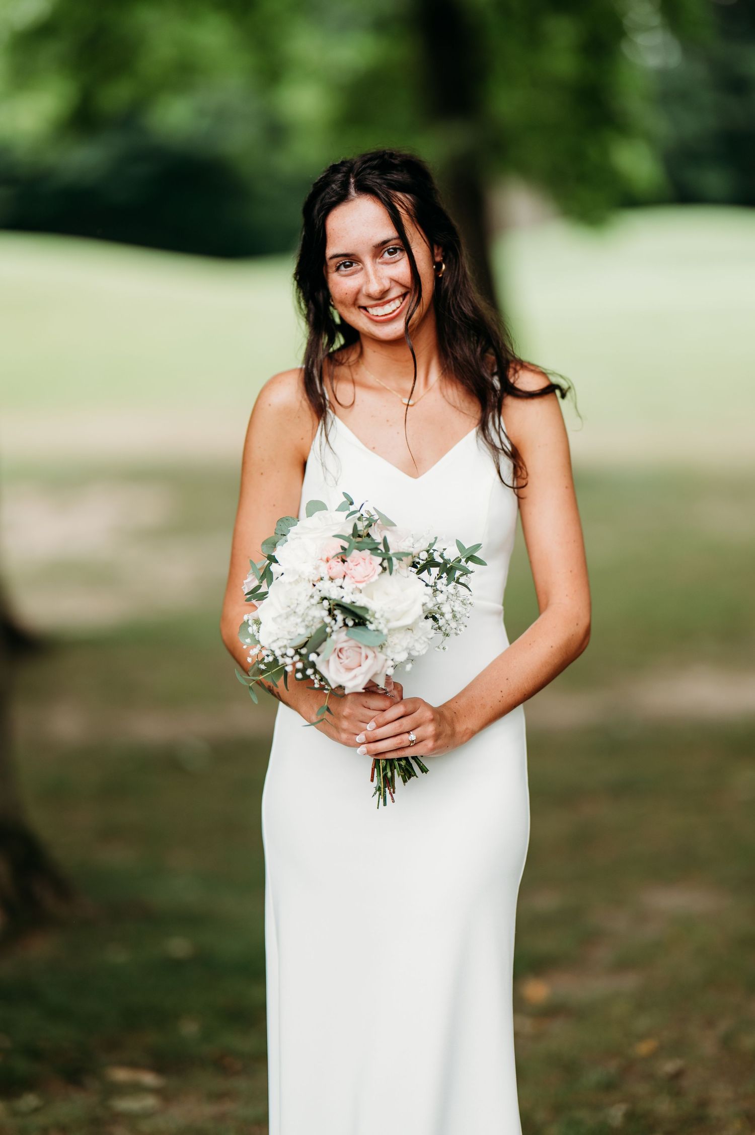 Bride in sleeveless white dress holds delicate pink and white floral bouquet in outdoor garden setting.