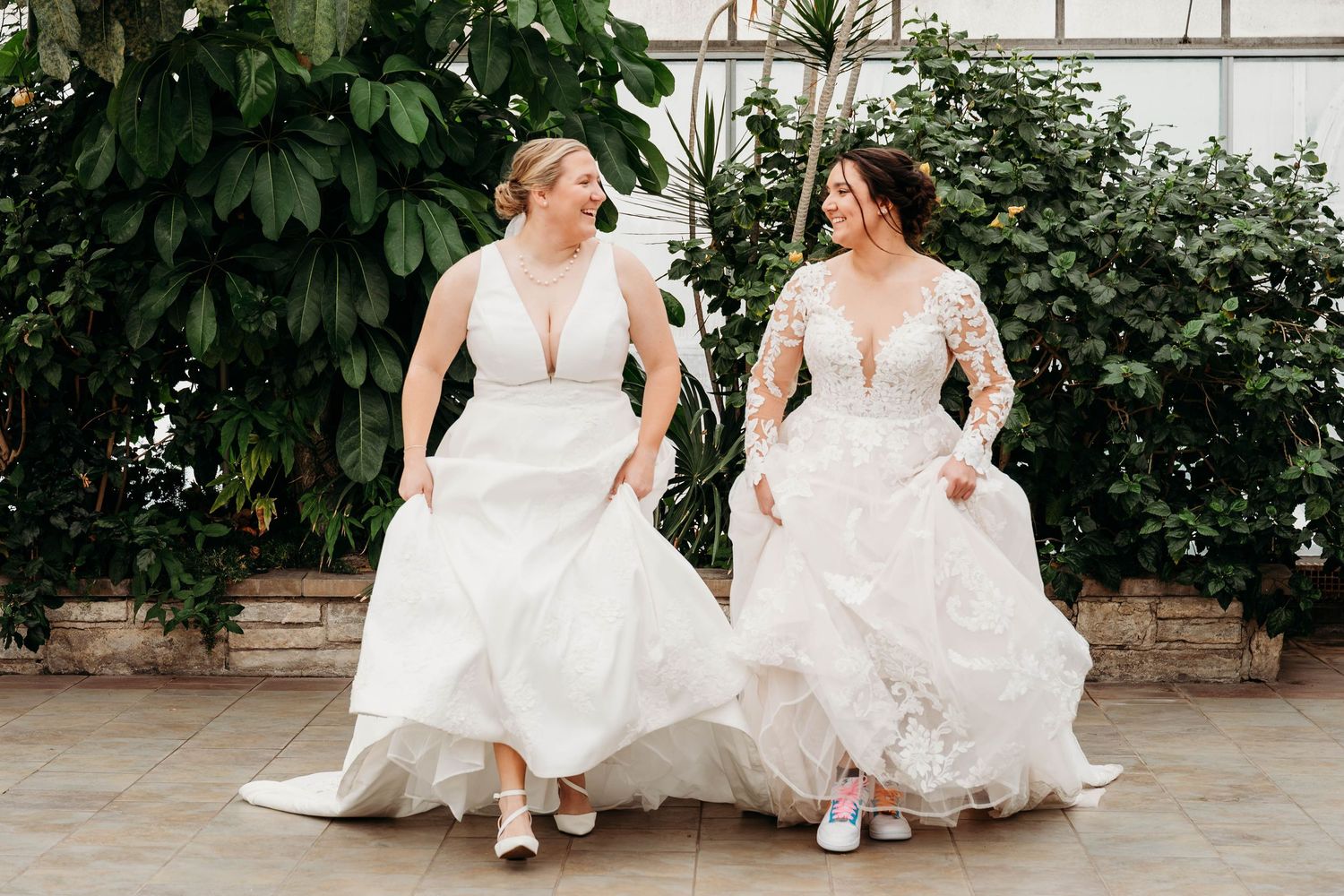 Two people in white wedding dresses sit together on stone steps surrounded by green foliage.