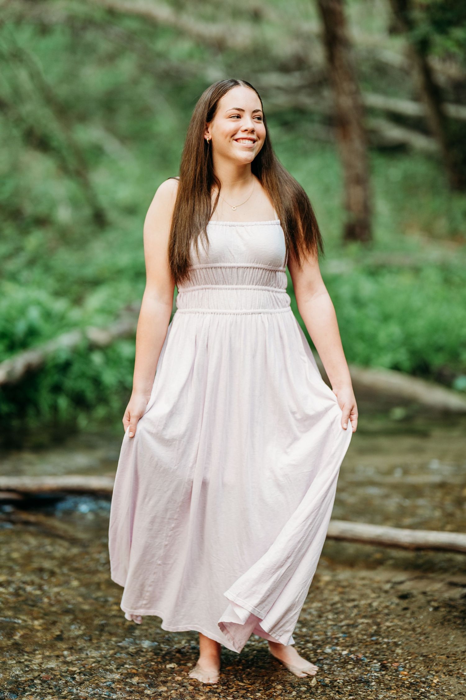 A person in a flowing white sundress poses playfully by a creek in a lush forest setting.