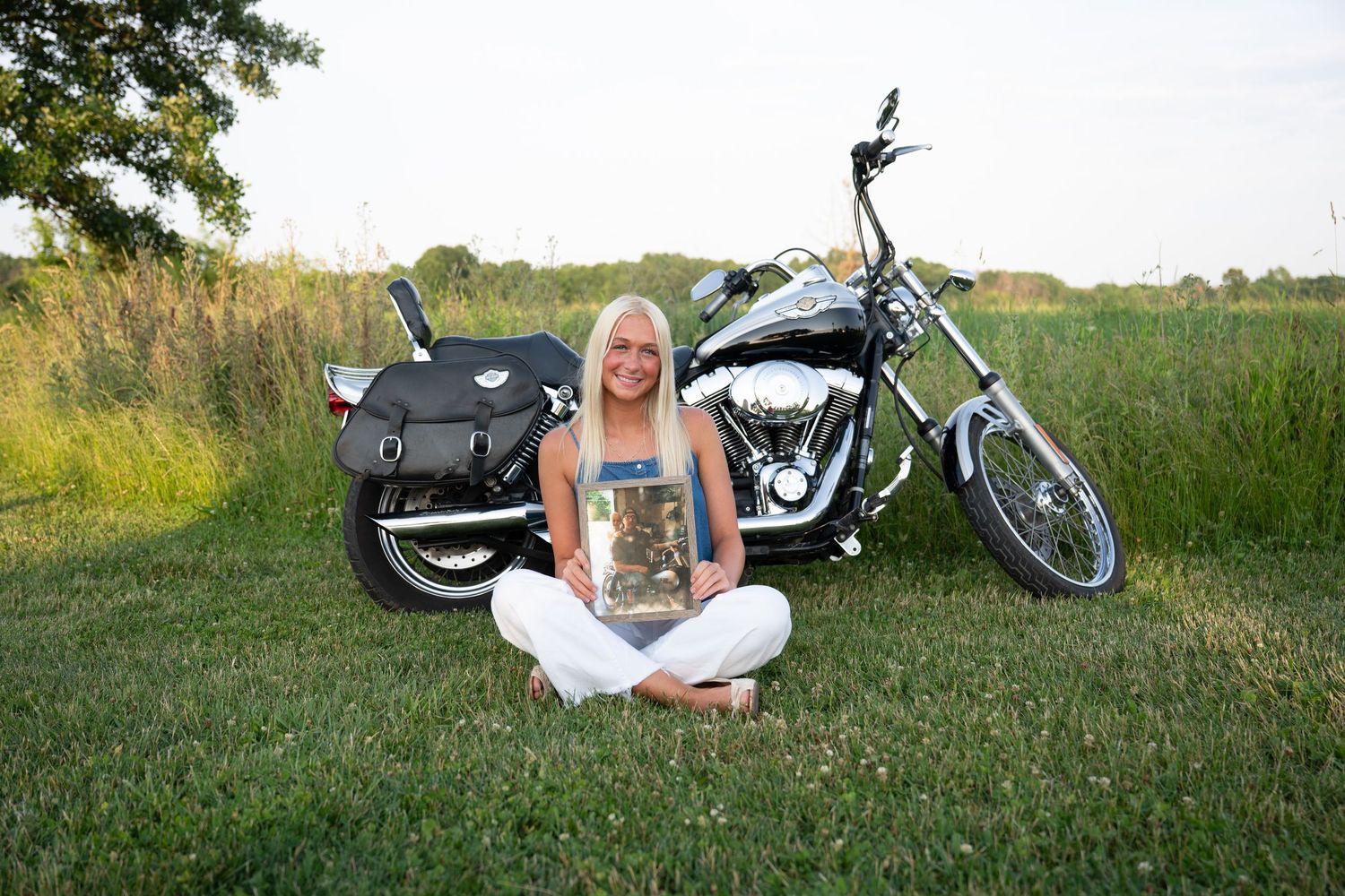 A motorcycle sits parked in a grassy field during golden hour with someone relaxing beside it.