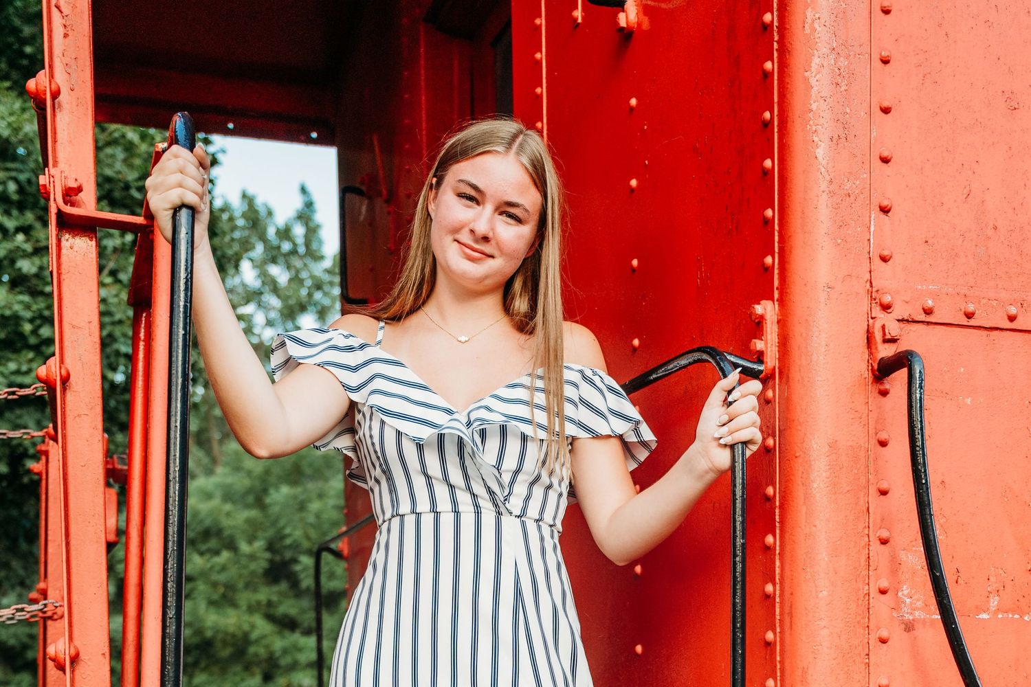 Woman in striped dress poses against red railroad car pillars on a sunny day.