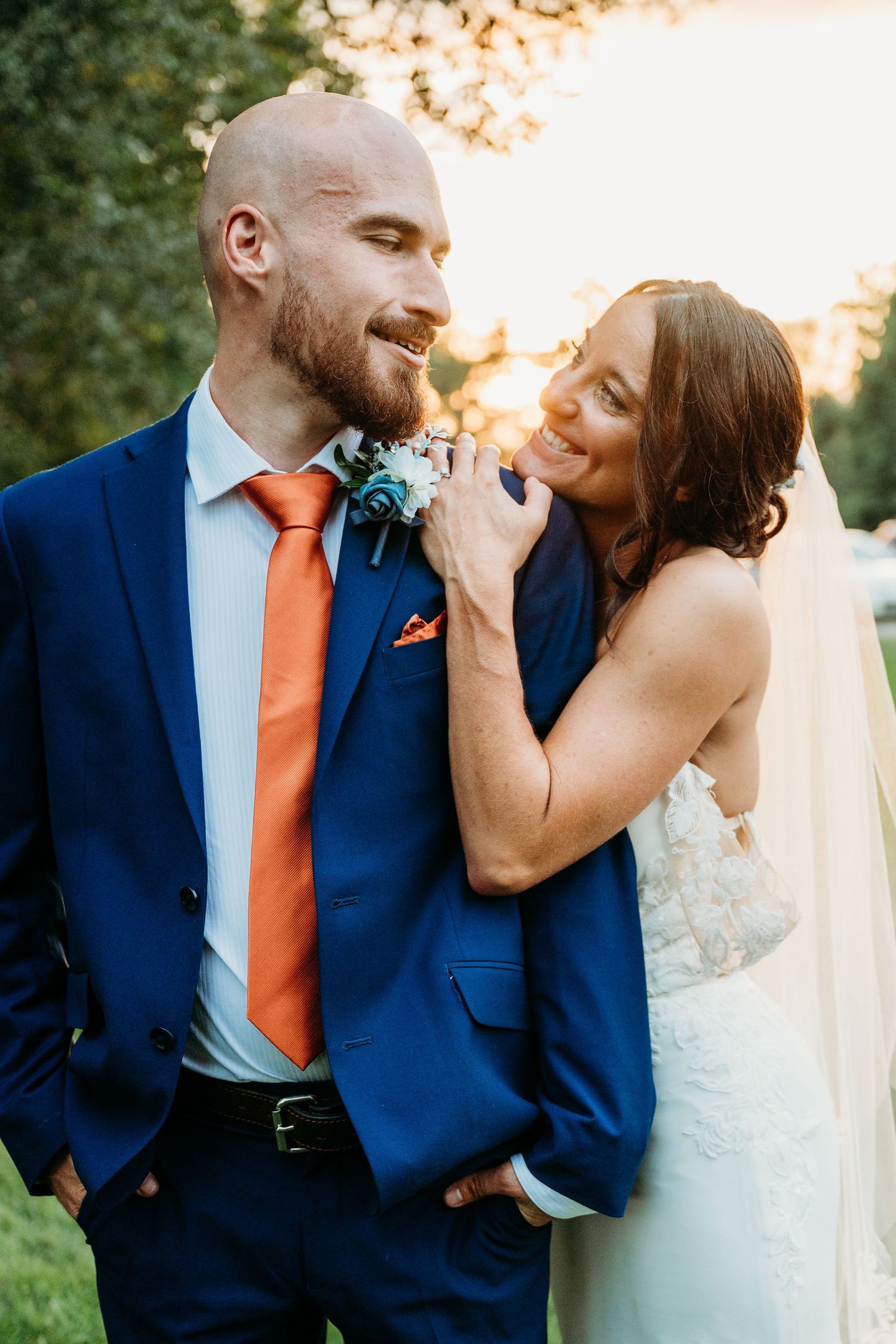Smiling bride in white gown and veil embraces bald groom in navy blue suit with orange tie at golden hour outdoor wedding.
