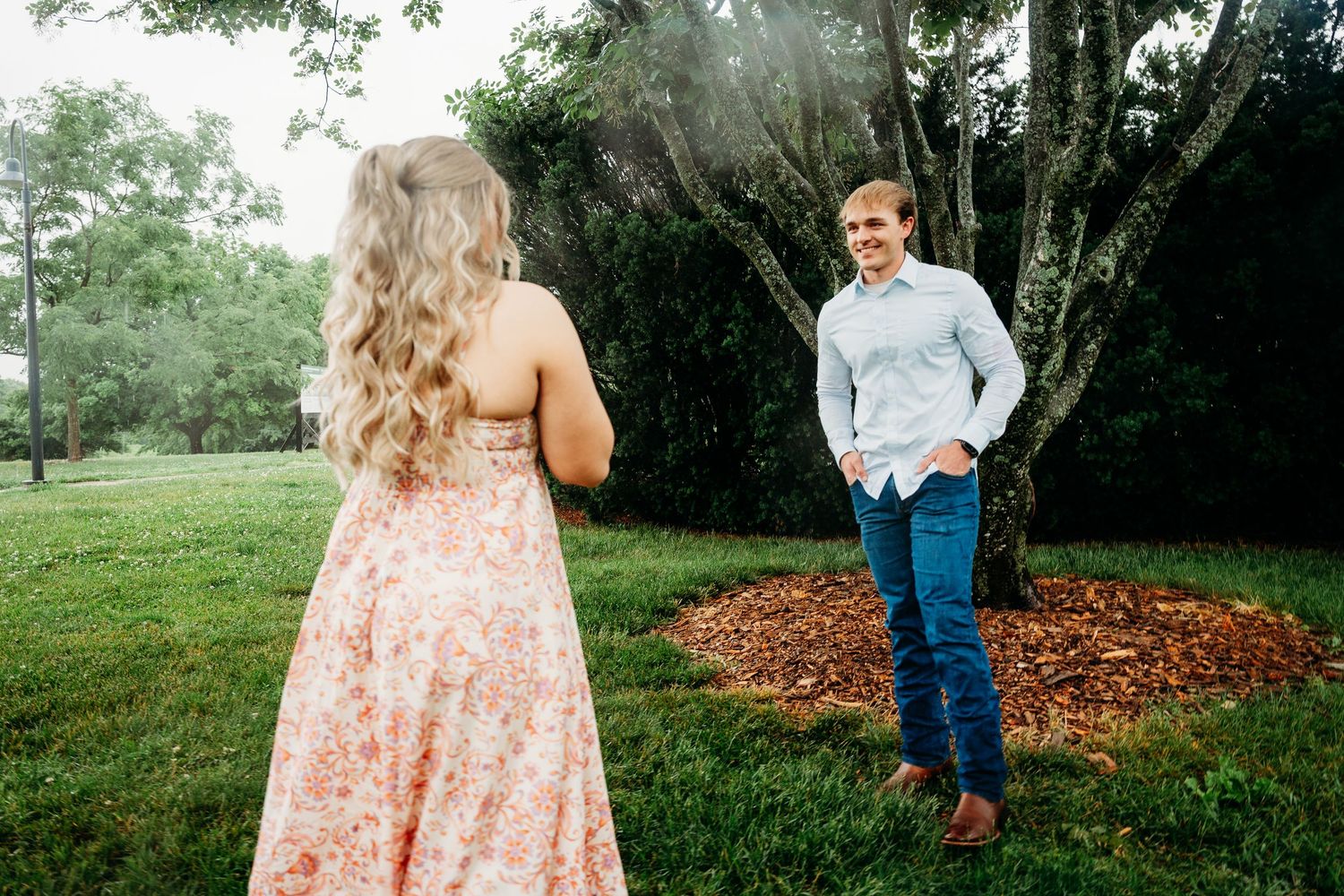 Couple standing outdoors on green grass near a large tree, woman in floral dress facing man in blue jeans and gray shirt.