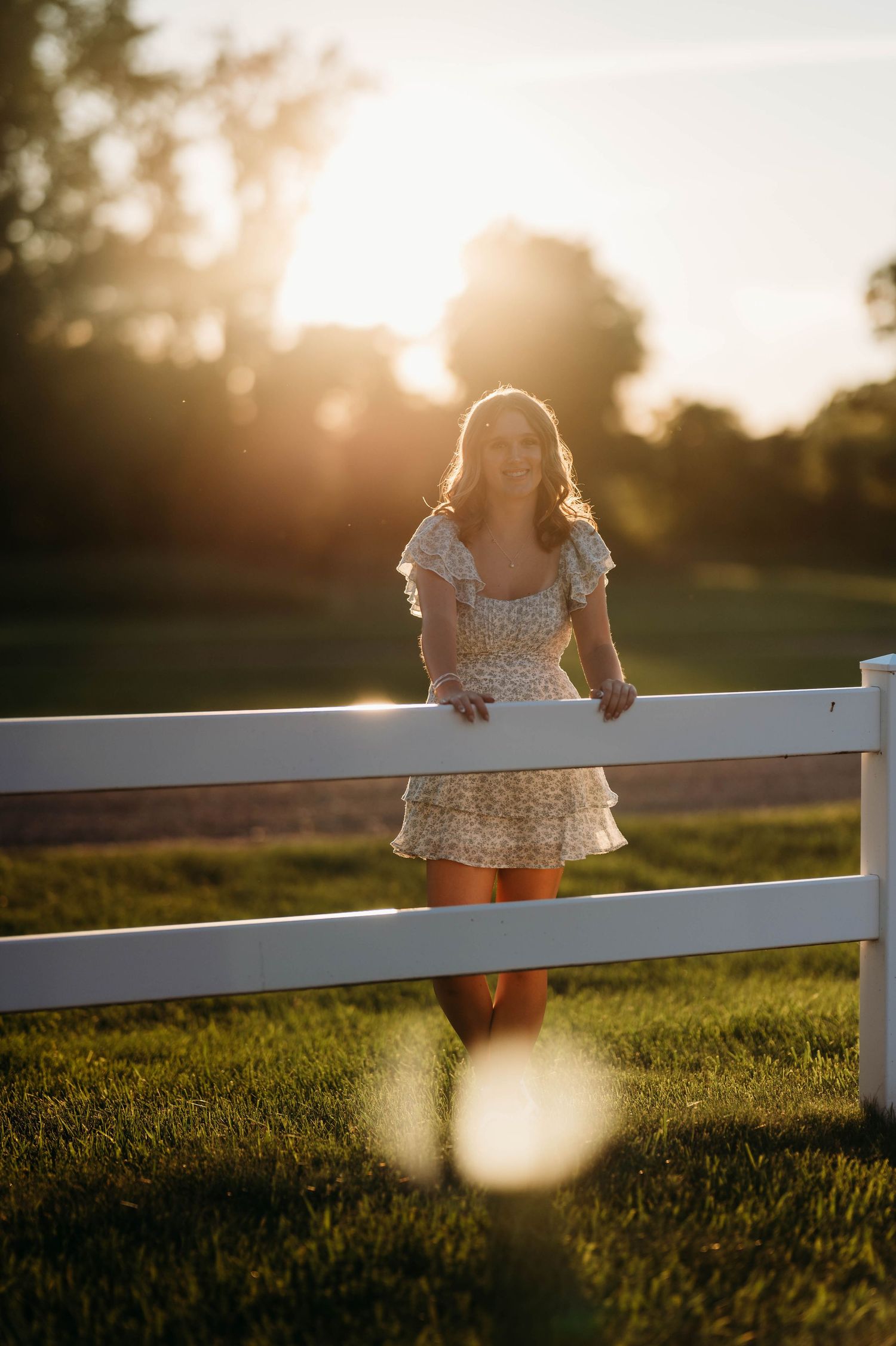 Young woman in floral dress leaning on white fence at golden hour sunset in green field.