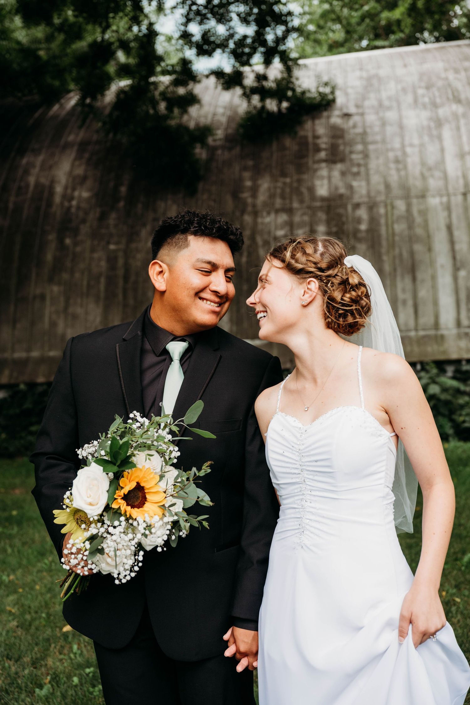 Newlywed couple smiling at each other, bride in white gown with veil, groom in black suit holding sunflower bouquet.
