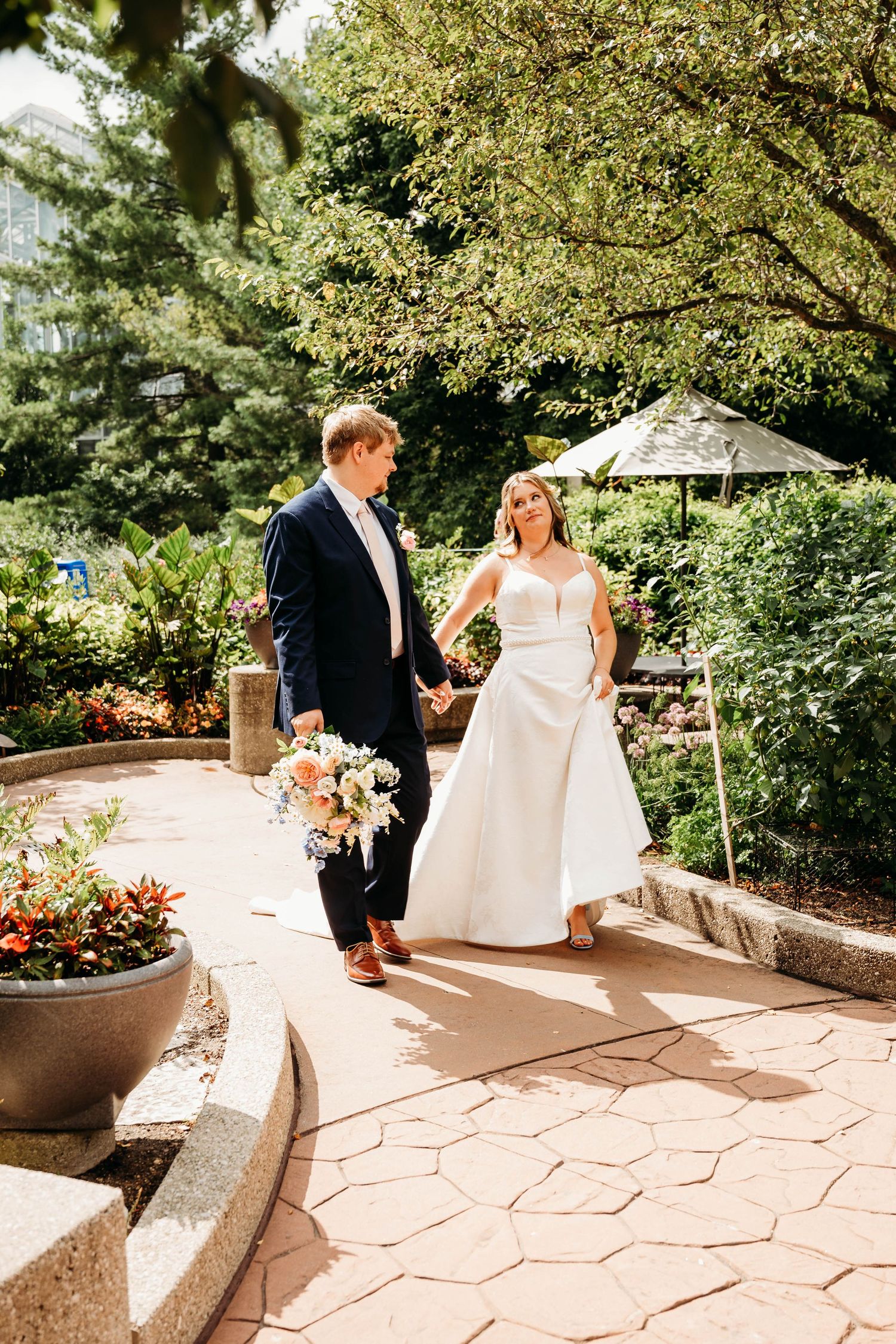 A bride with a cane and groom in navy suit walk hand-in-hand along a garden path, surrounded by lush greenery.