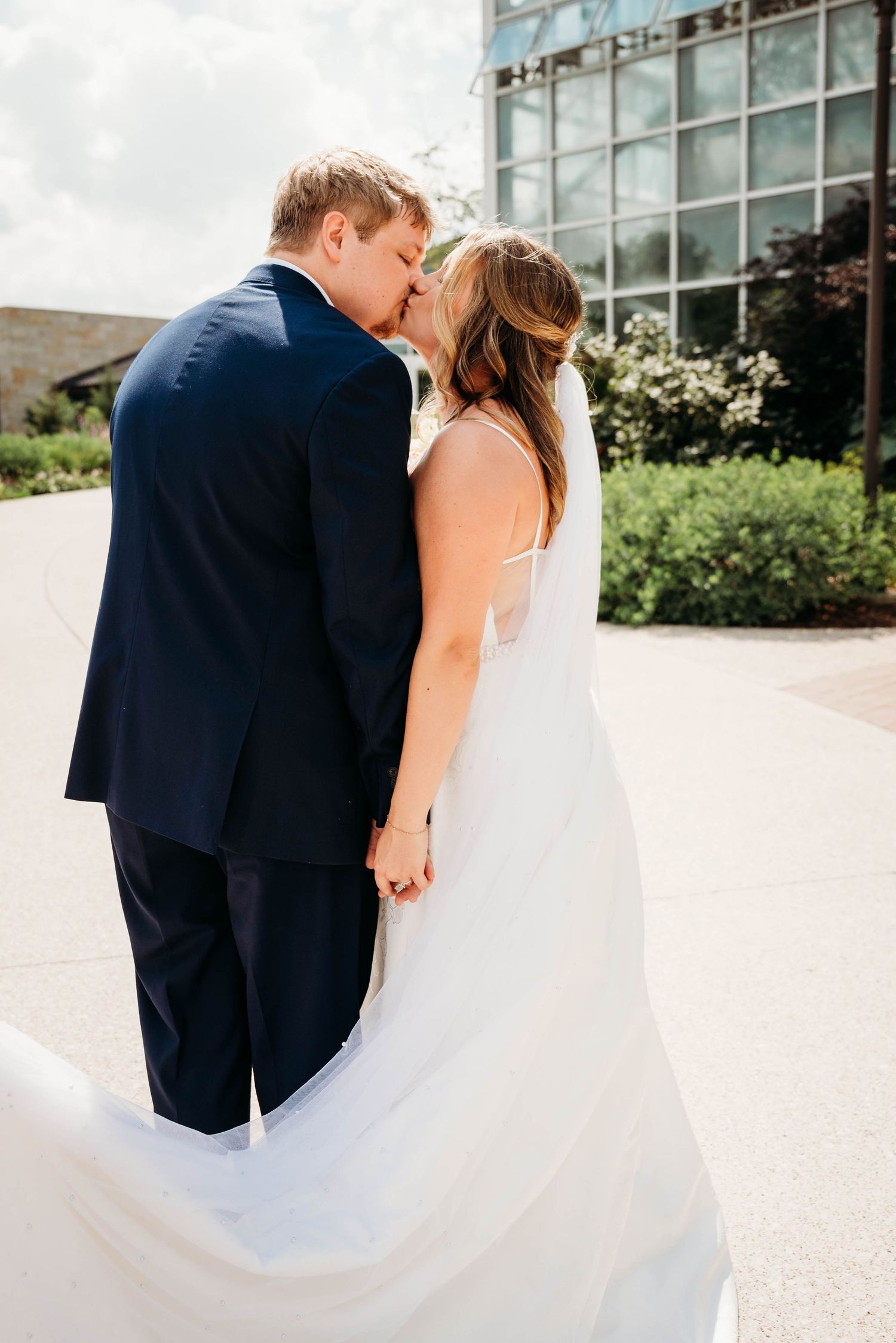 Bride and groom share a kiss outdoors, holding hands, wearing a white gown and navy suit on their wedding day.