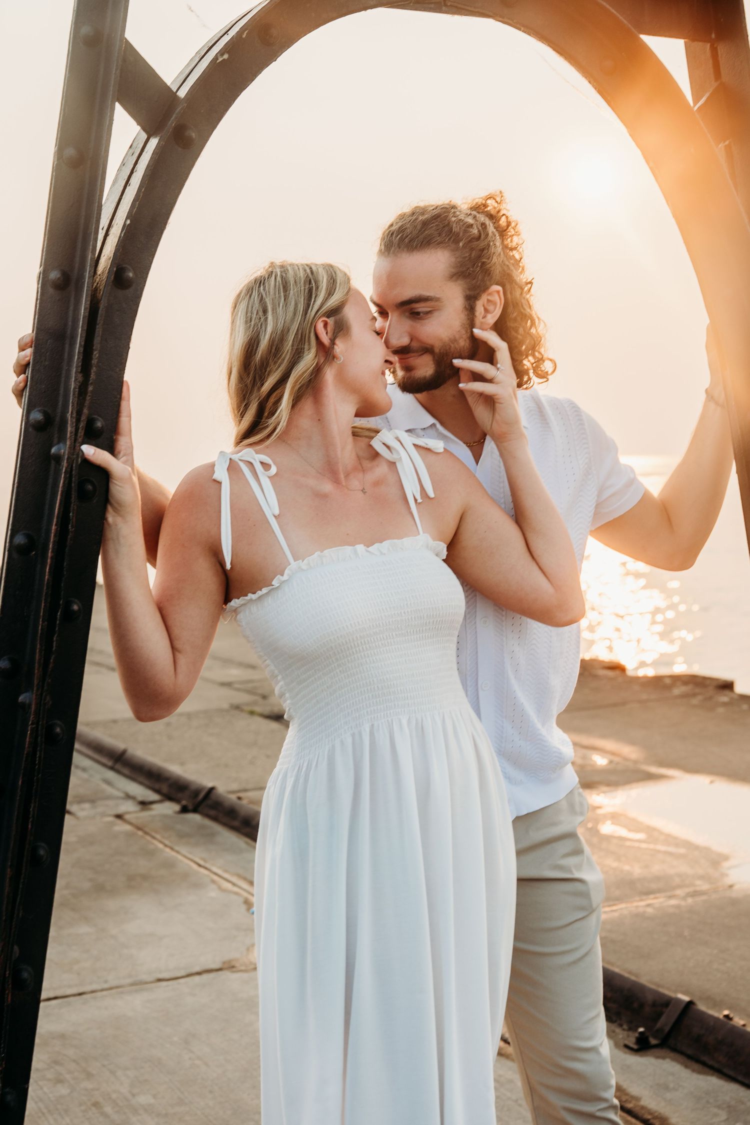 Couple sharing a romantic almost-kiss at sunset by the sea, woman in white dress, man in white shirt near iron arch.