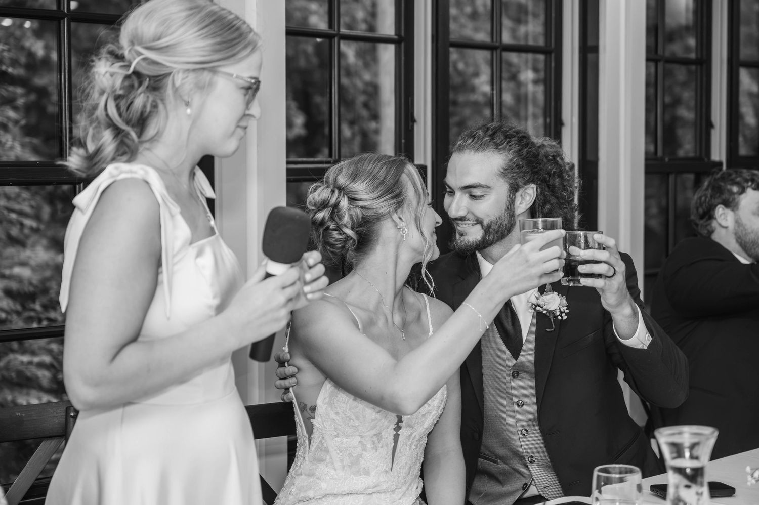 Black and white photo of bride and groom toasting drinks with bridesmaid holding microphone at wedding reception.