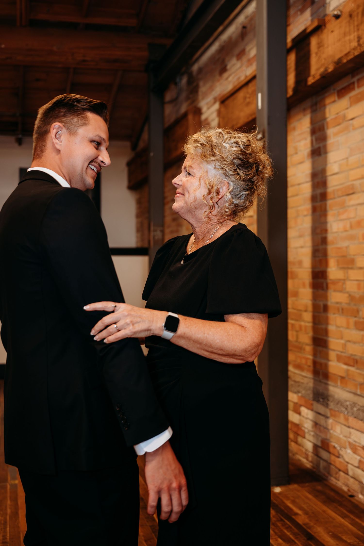 Smiling groom in tuxedo shares a warm moment with older woman in black dress at wedding reception.