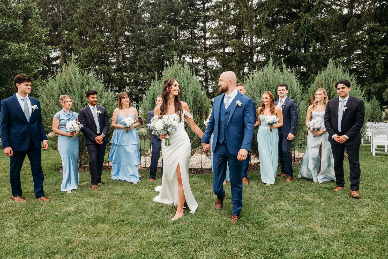 Wedding party poses outdoors on green lawn, bride and groom walk hand-in-hand flanked by bridesmaids in blue and groomsmen.