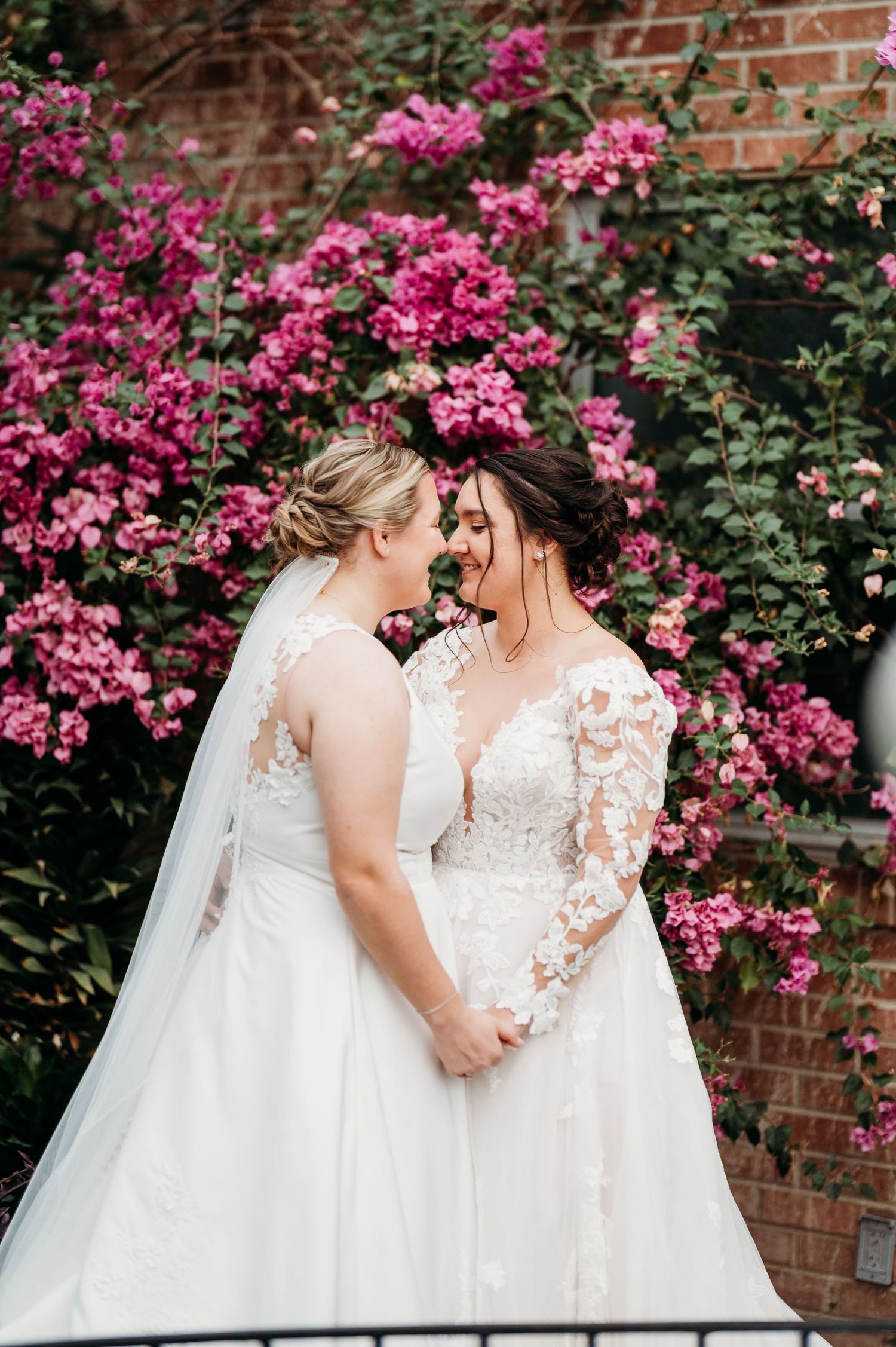 Two wedding dresses with delicate lace details stand together against a backdrop of vibrant pink flowers and brick wall.