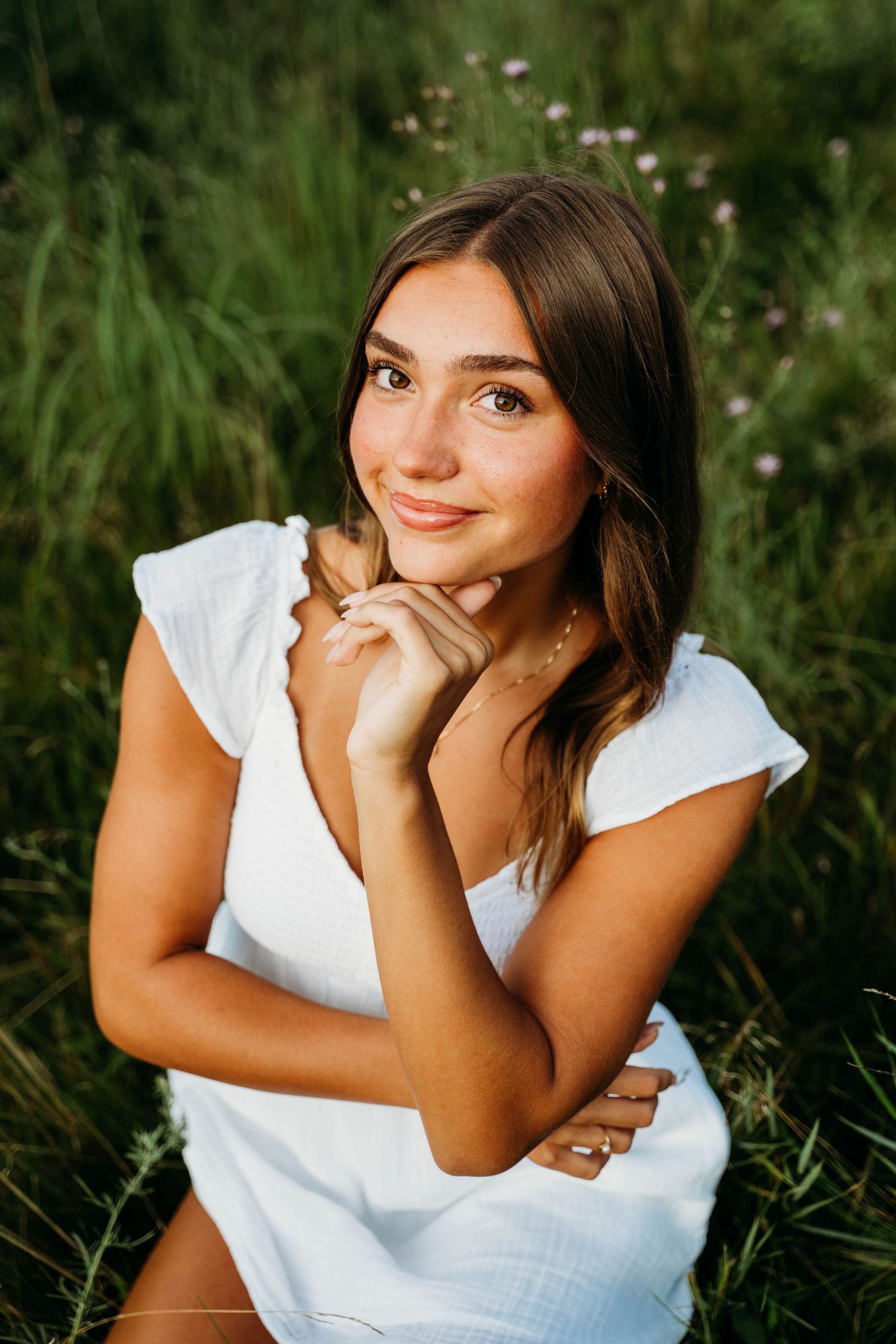 Portrait of someone in a white summer dress sitting in a grassy meadow with wildflowers at sunset.