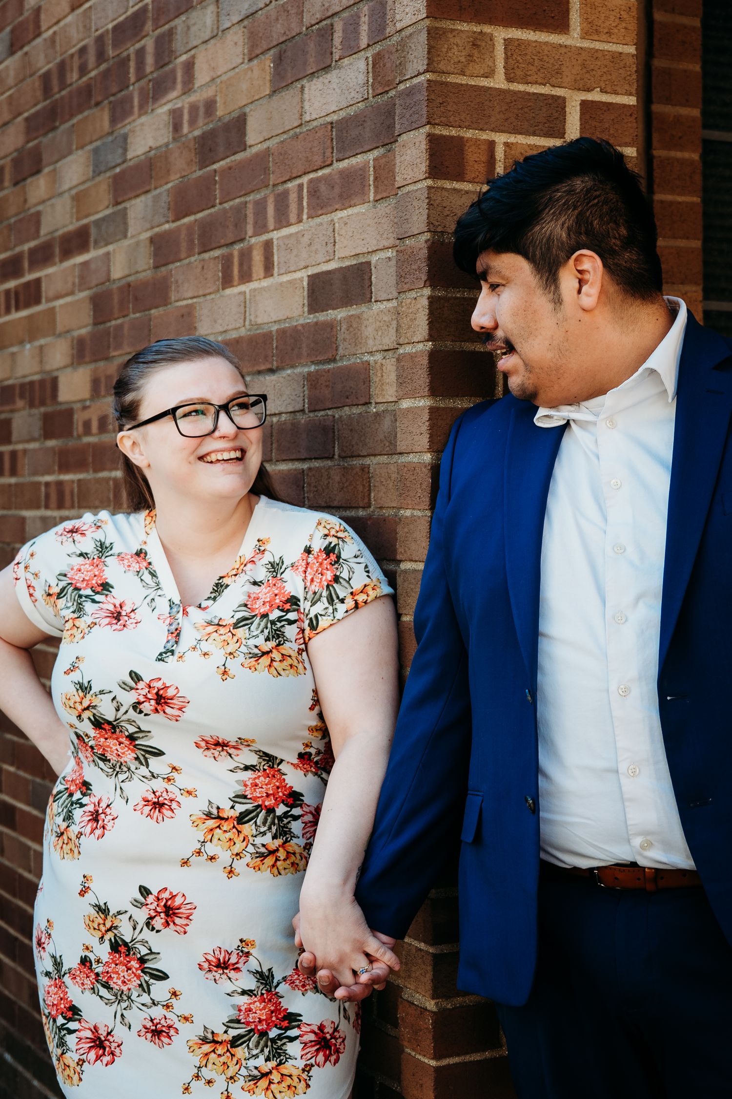 A couple stands holding hands and smiling at each other against a brick wall, she in a floral dress and he in a blue blazer.