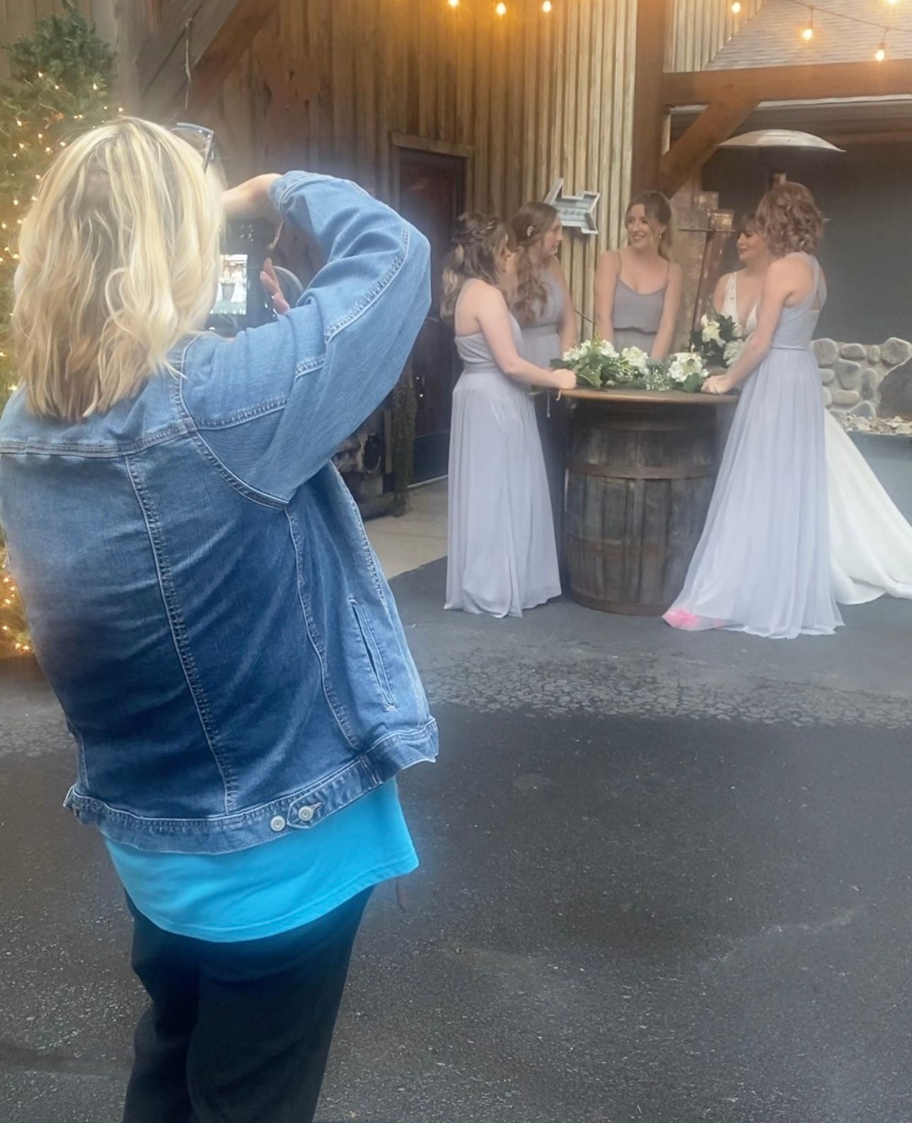 Photographer in denim jacket captures a wedding moment at a rustic barn venue with string lights and floral decor.