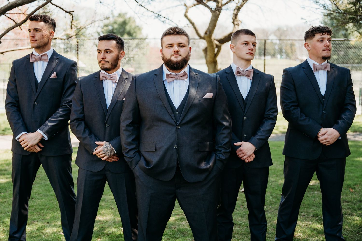 A group of five groomsmen in matching navy suits stand together on grass during a wedding photo session.