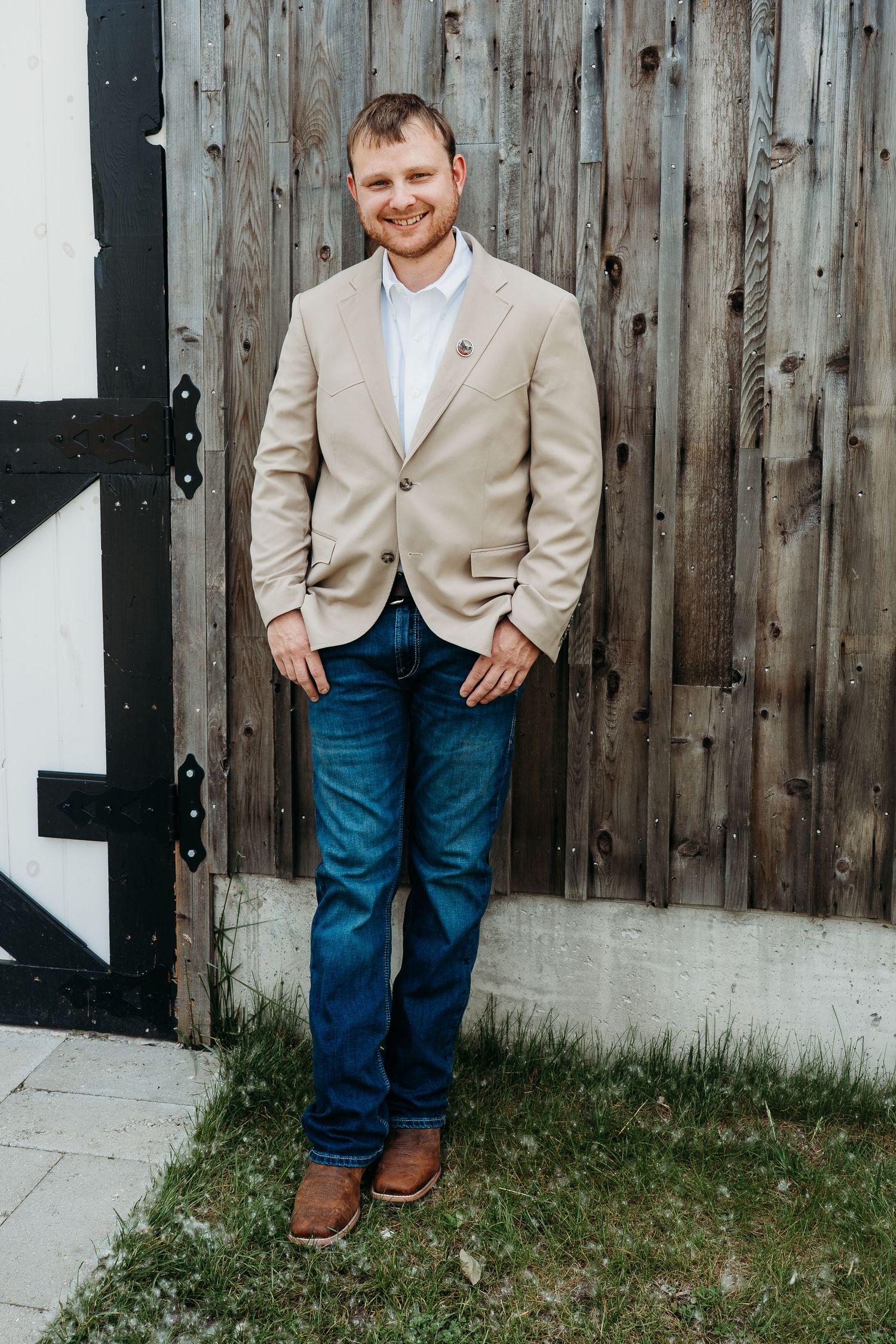 A figure in a beige blazer and blue jeans stands against a rustic wooden fence backdrop.