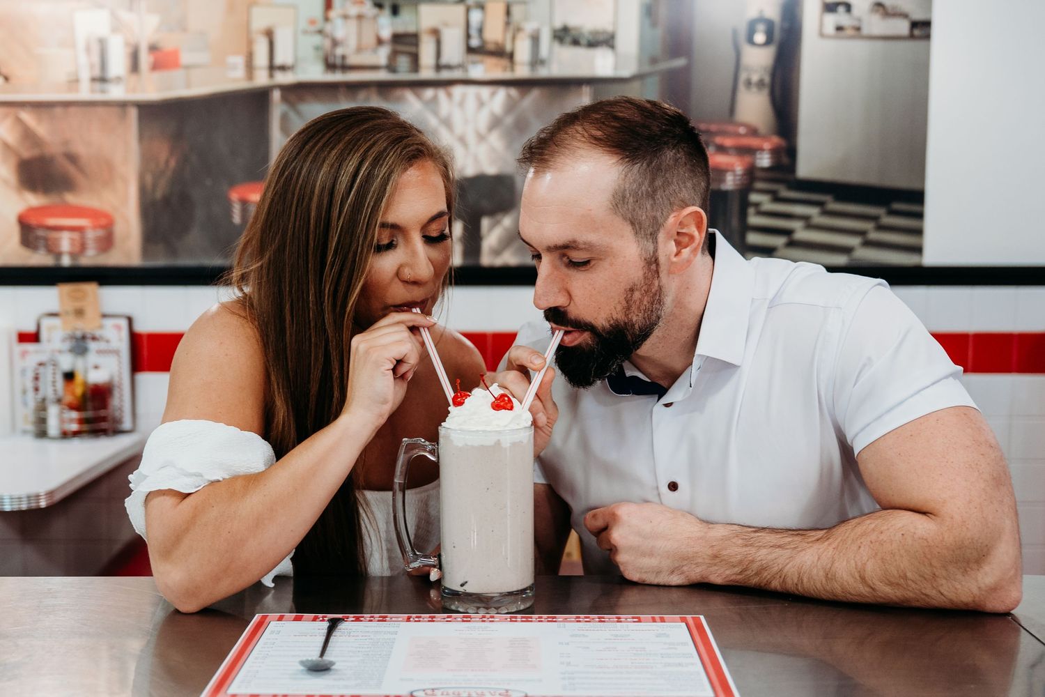 A romantic couple leans in close while sharing a milkshake at a diner.