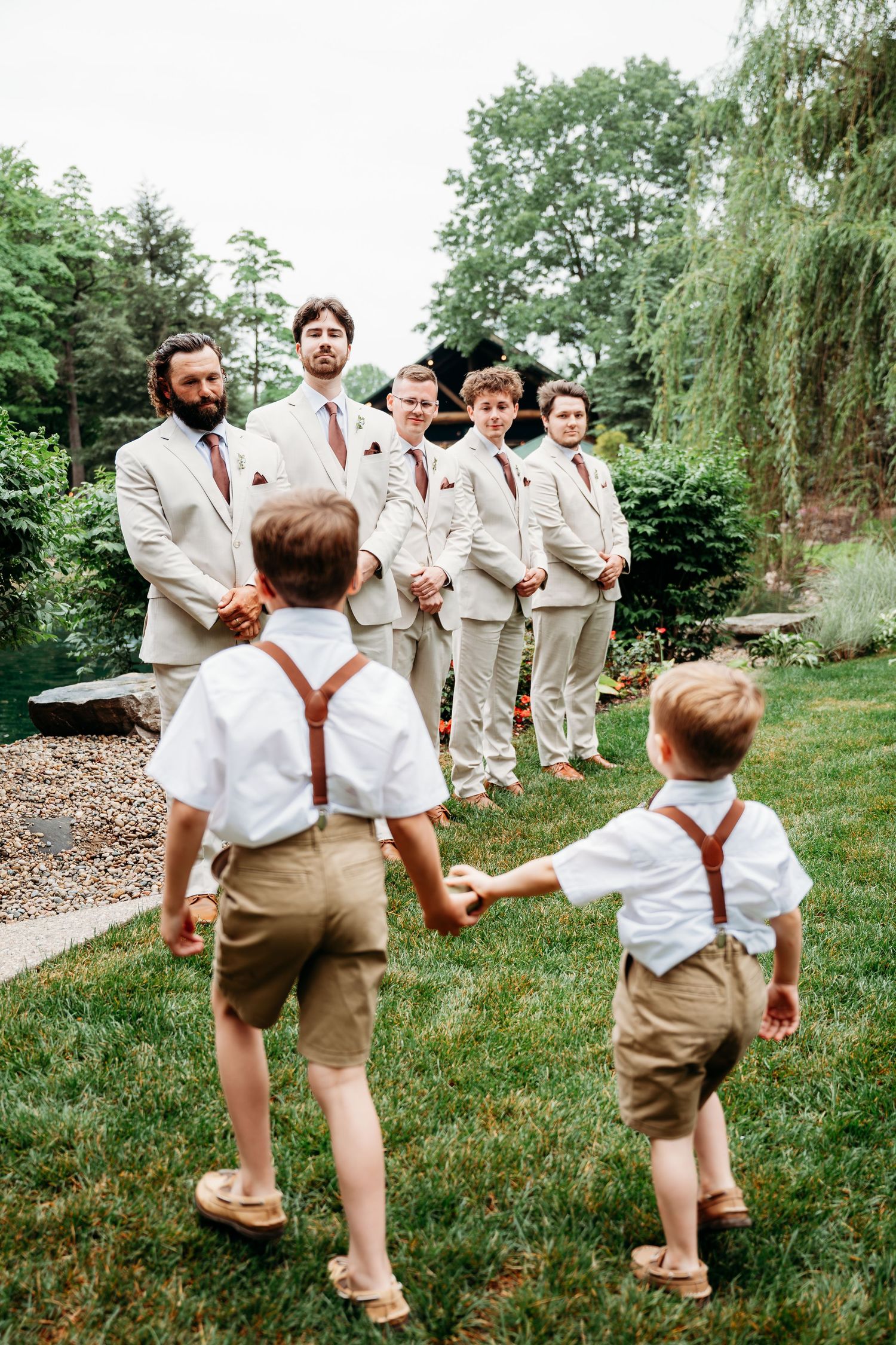 Two ring bearers in suspenders approach groomsmen in tan suits at an outdoor wedding ceremony.