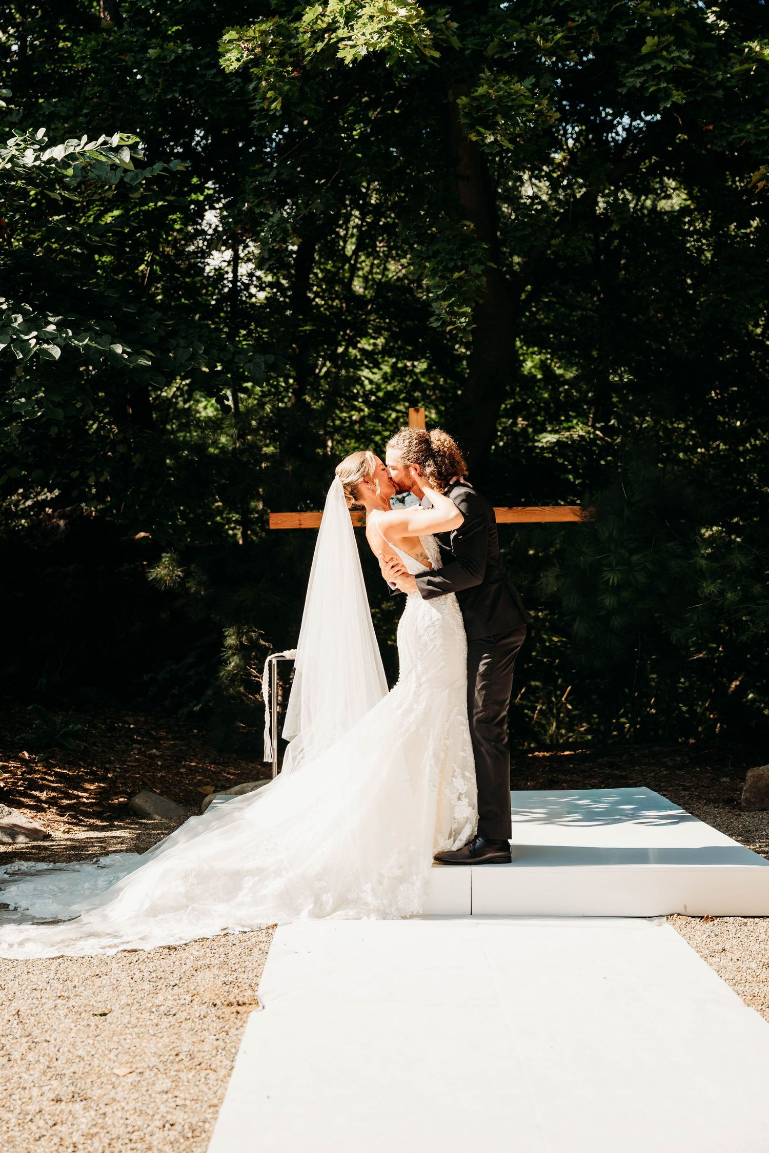 Bride and groom share first kiss at outdoor wedding ceremony surrounded by lush green trees on sunny day.