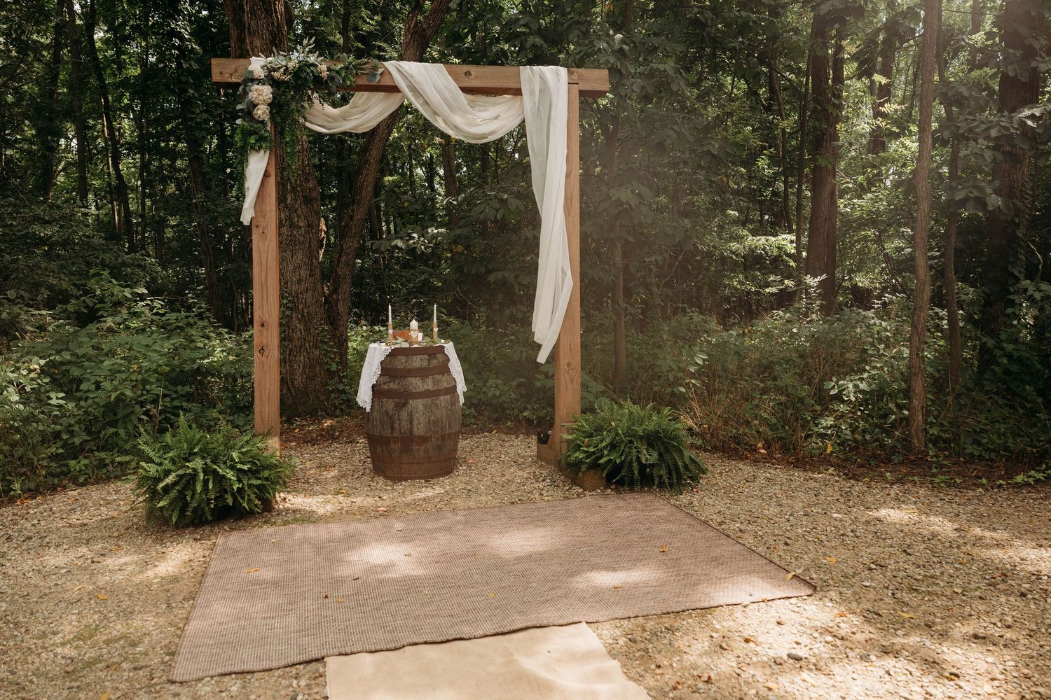 A rustic wooden wedding arch with white fabric draping stands in a forest clearing with potted plants at its base.