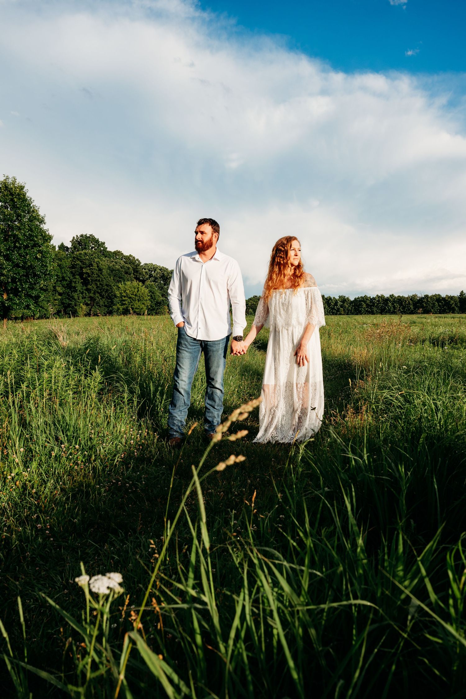 Couple holding hands in a green meadow, man in jeans and white shirt, woman in white lace dress, under blue sky.