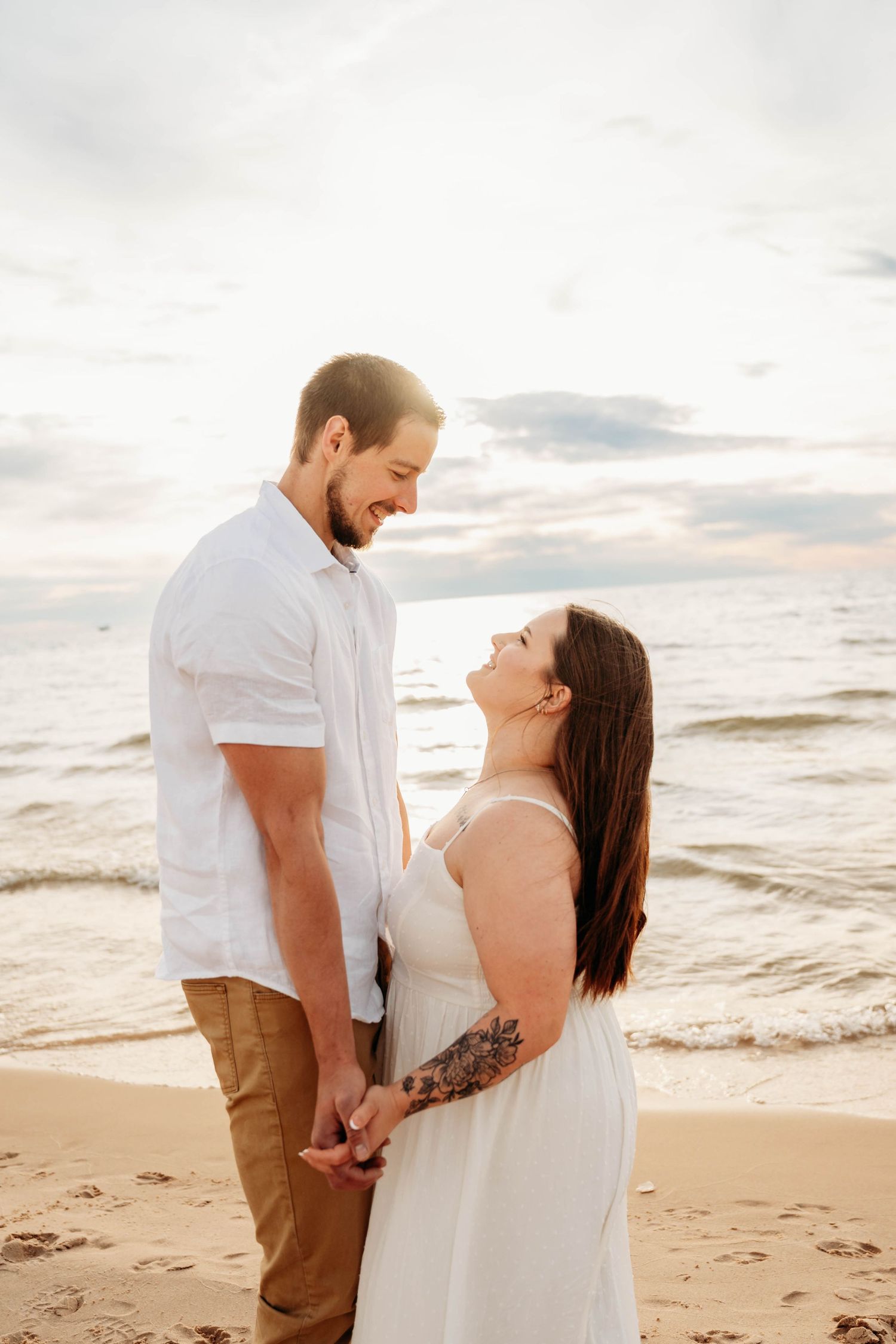 Romantic couple holding hands and smiling at each other on a beach at golden hour, woman in white dress.