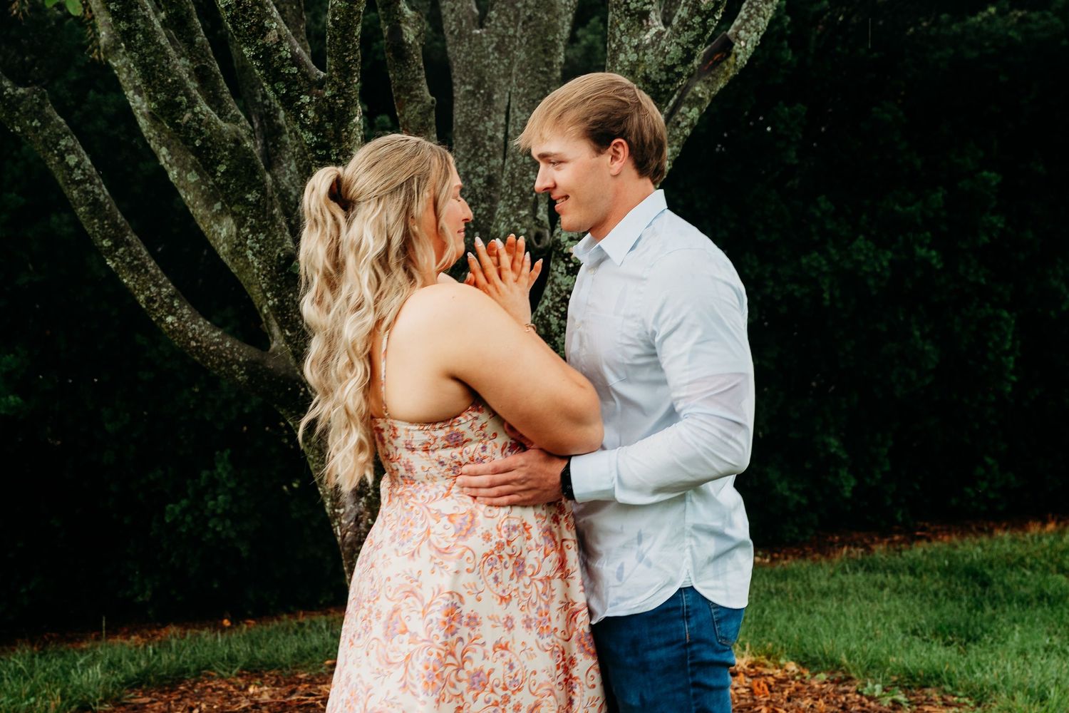 Couple embracing outdoors near a large tree, woman in floral dress showing engagement ring, man in white shirt and jeans.