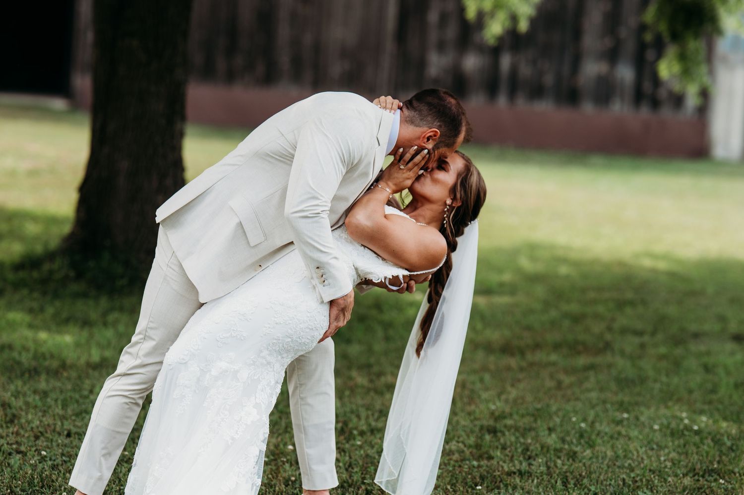 A romantic wedding couple embraces in a dramatic dip pose on a grassy lawn surrounded by trees.