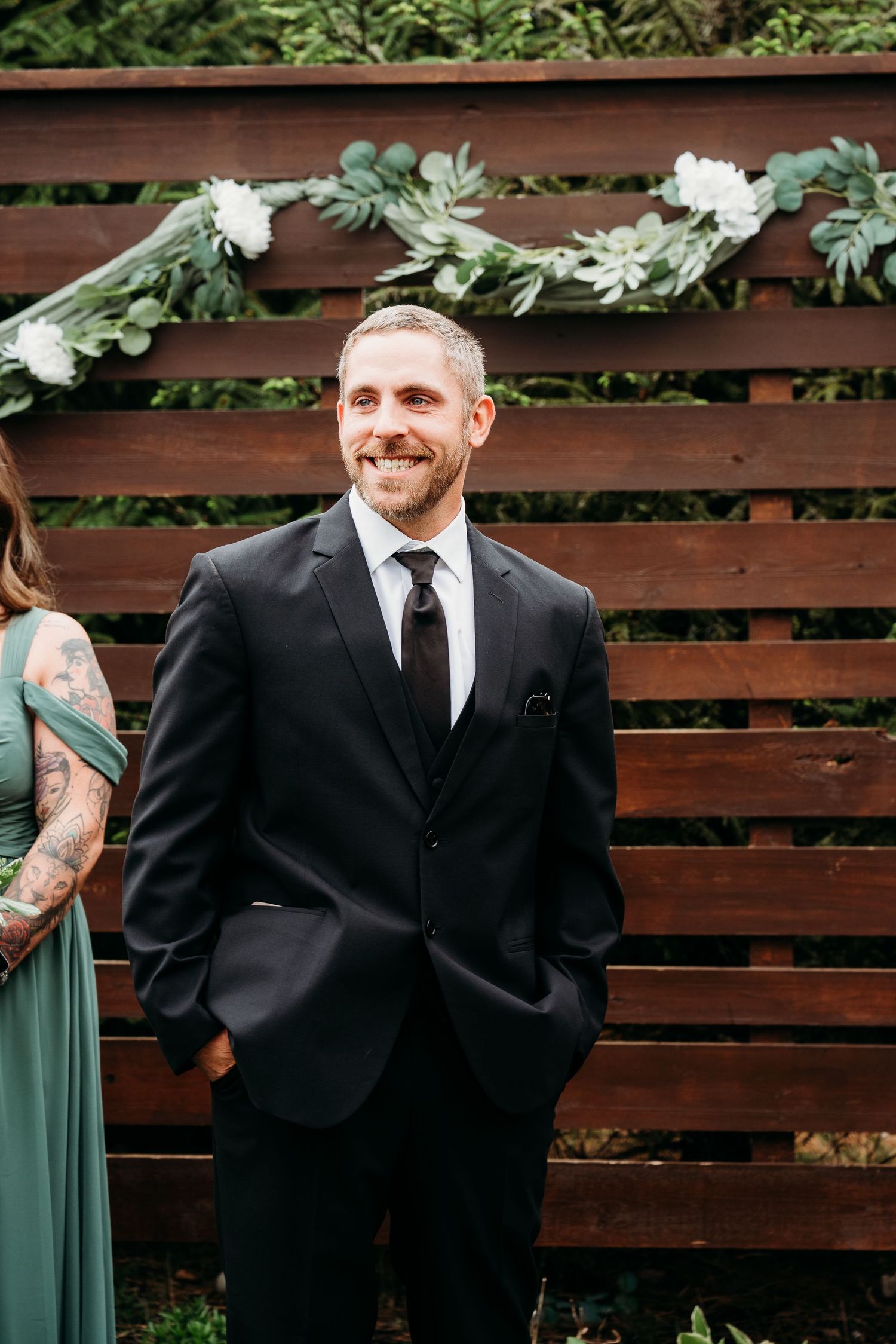 Wedding party members in formal attire stand against a rustic wooden fence backdrop during an outdoor celebration.