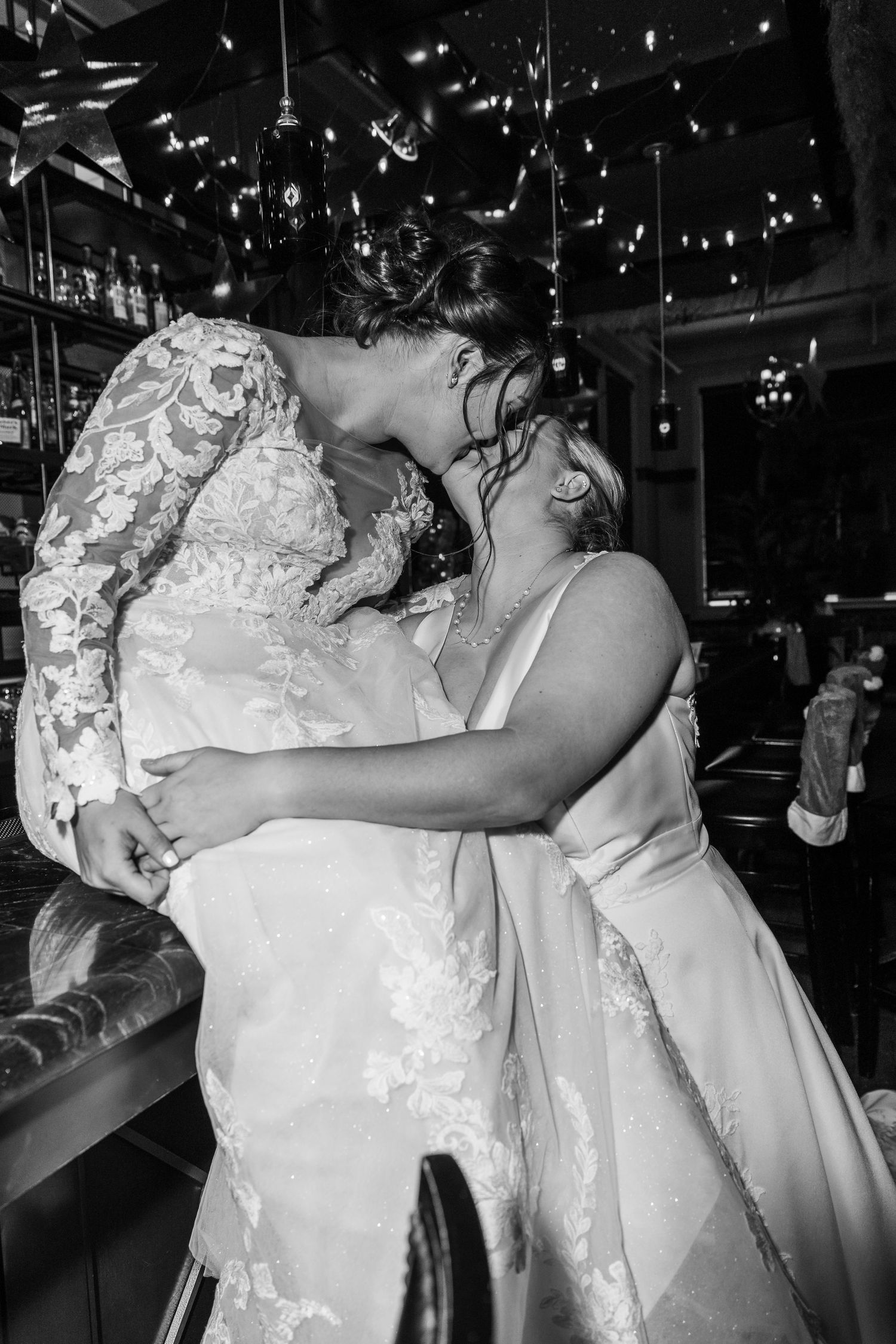 Two people in white wedding attire embrace during a celebration under twinkling lights.