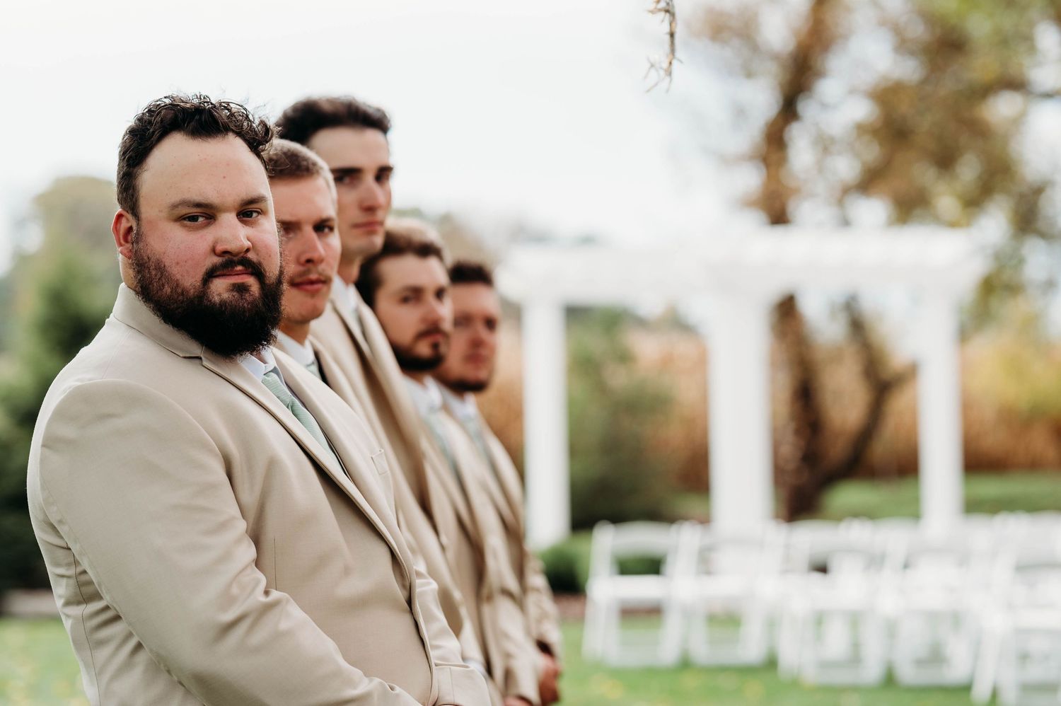 A row of groomsmen in beige suits stand together at an outdoor wedding ceremony setup with white chairs.