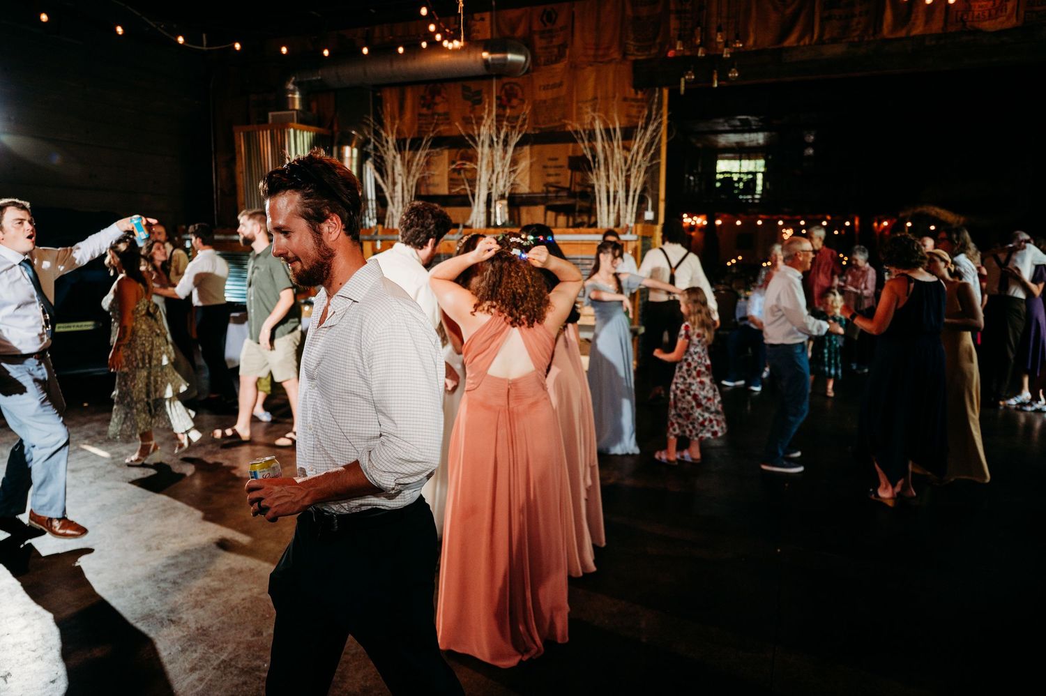 Guests dancing at a lively wedding reception in a rustic venue, with a woman in a coral bridesmaid dress.