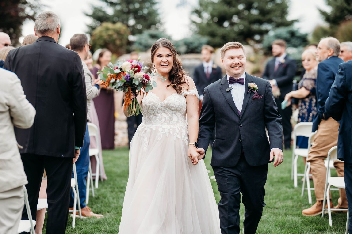 Newlywed couple walking down outdoor wedding aisle, bride in white gown holding bouquet, groom in navy suit with bow tie.