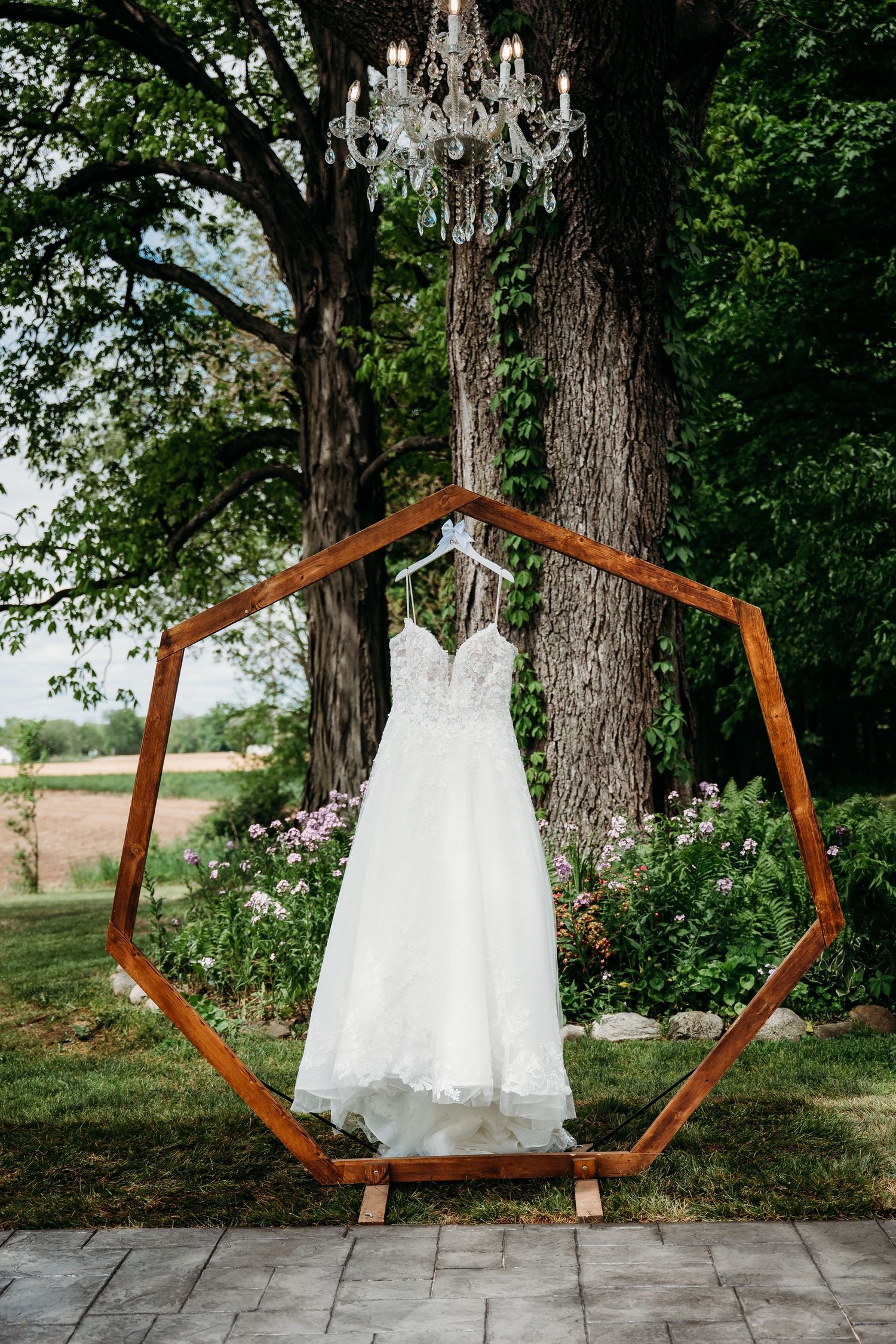 A white wedding dress hangs from an octagonal copper frame with a crystal chandelier above and trees in the background.