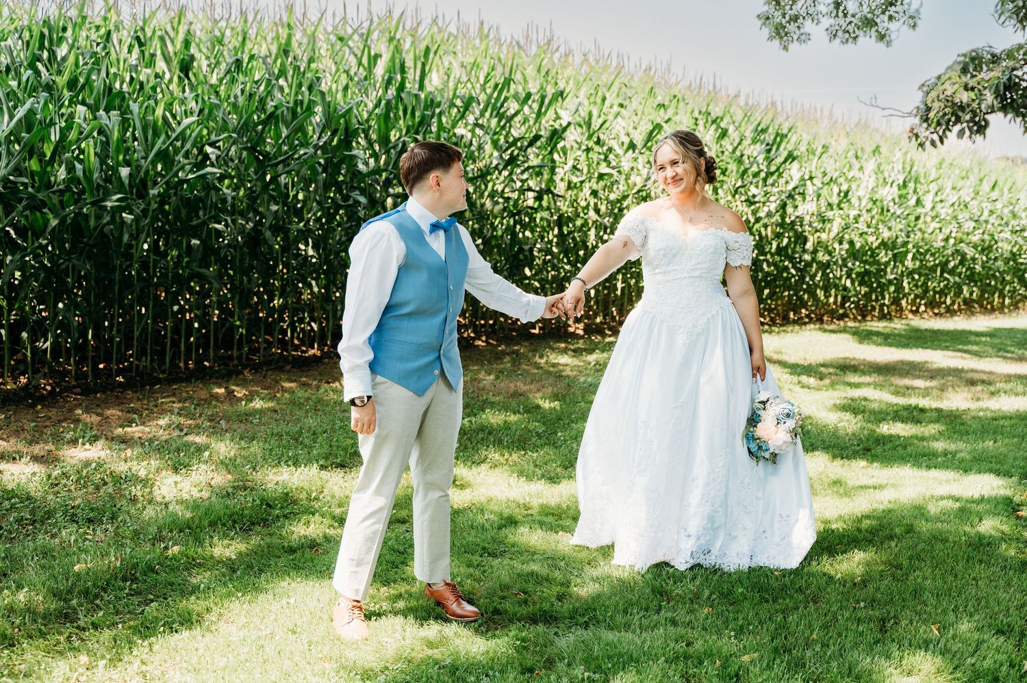 Couple in formal wedding attire walk on green grass path with cornfield and trees in the background on sunny day.