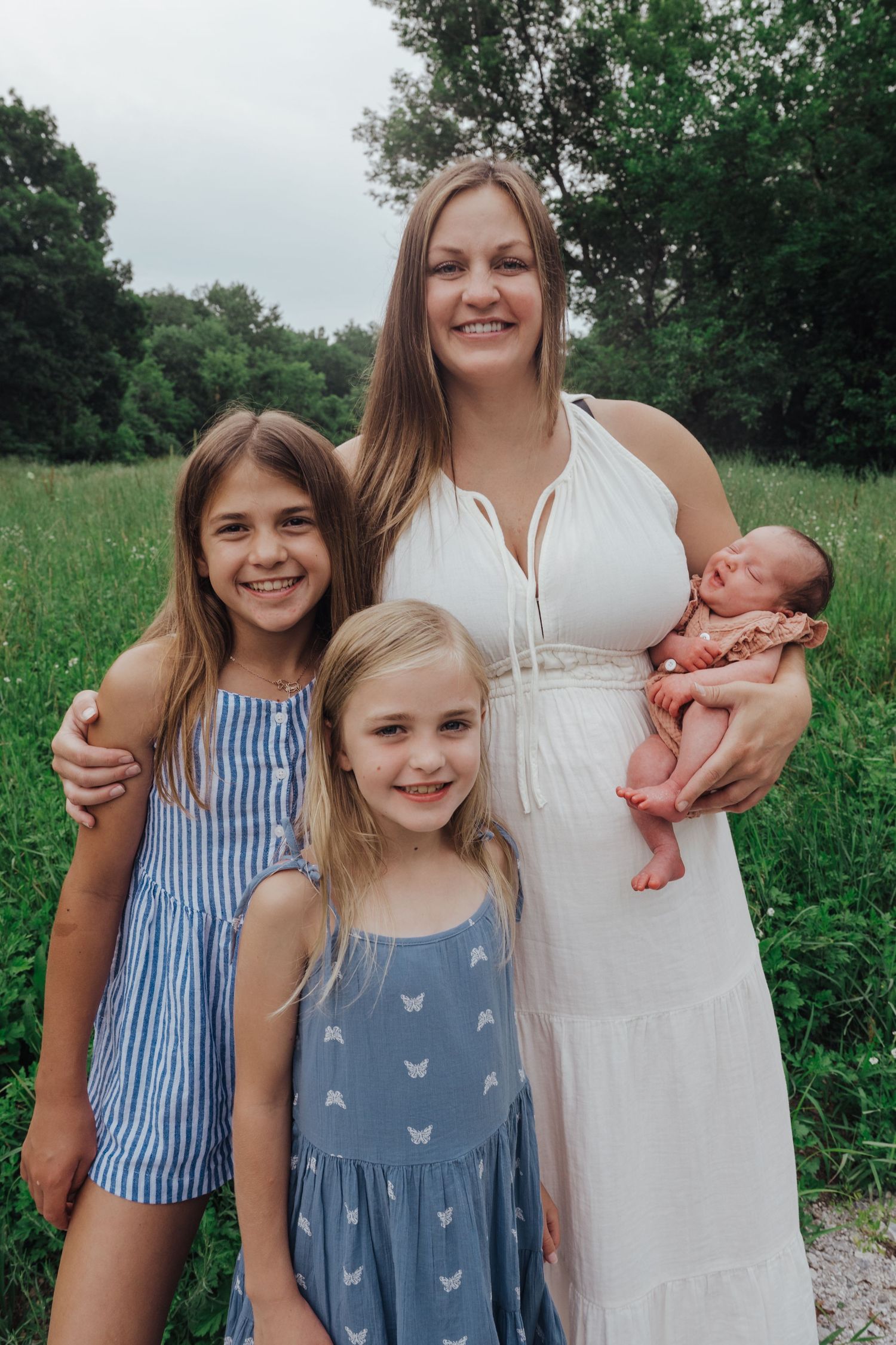 A family poses outdoors in a grassy field wearing summer dresses with an infant in arms.